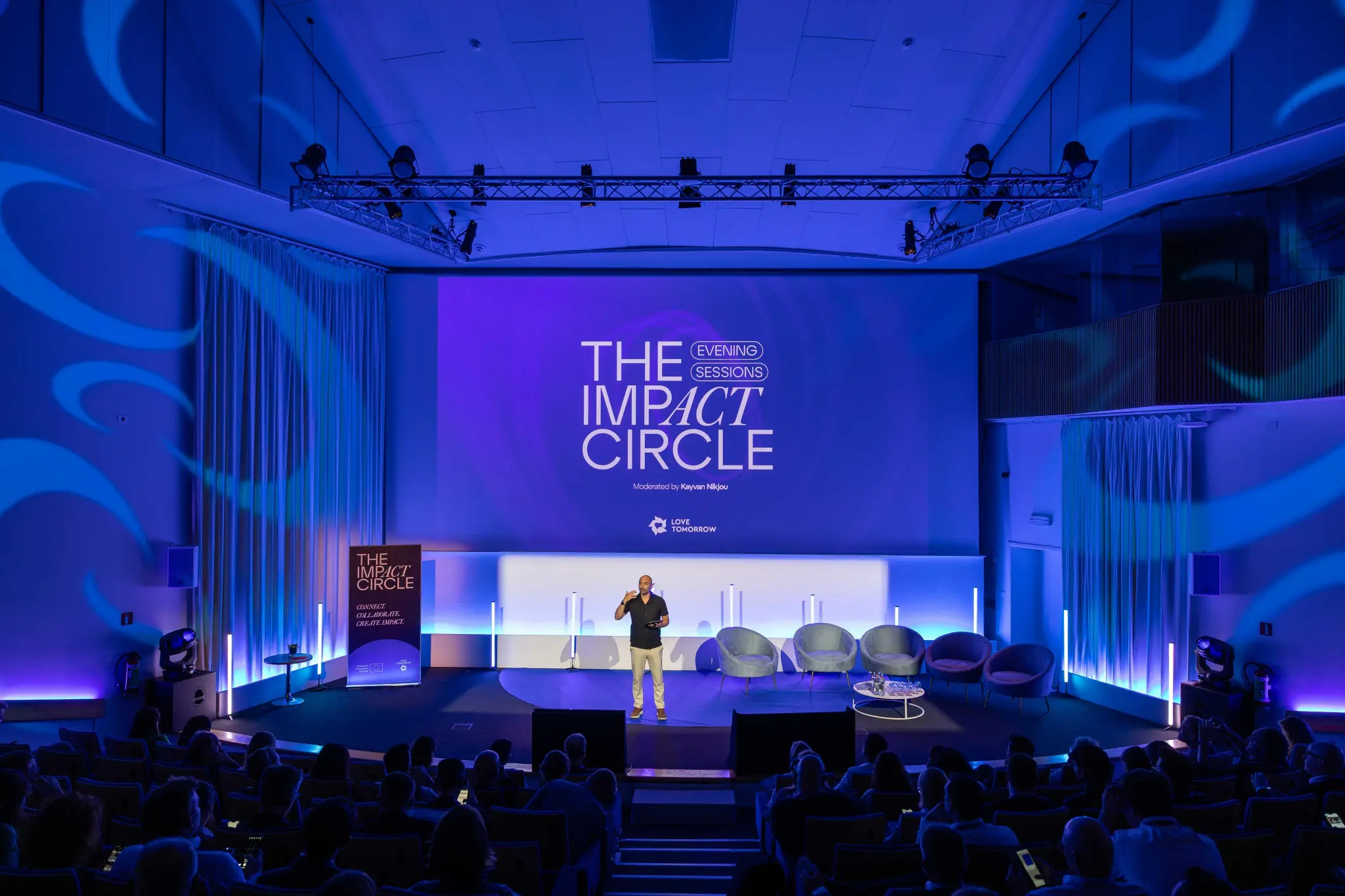 Man speaking on stage in a blue-lit auditorium with a screen displaying 'The Impact Circle Evening Sessions' and an audience in the foreground at the Love Tomorrow Summit