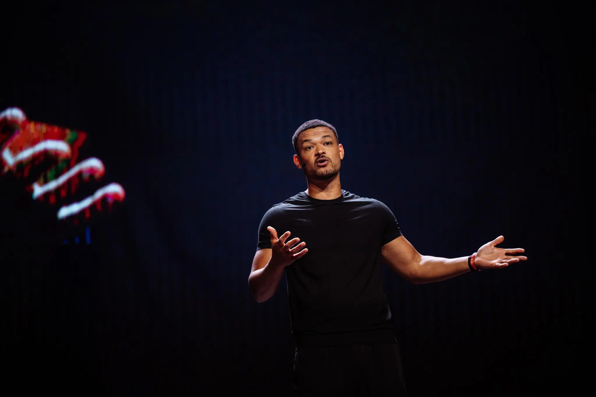 Steven Bartlett in black shirt speaking on stage with a dark background and a colorful abstract shape on the left at the Love Tomorrow Summit