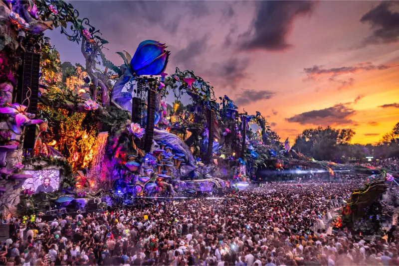 Large crowd attending an outdoor music festival at sunset with an elaborate, colorful stage decorated with oversized flowers and vegetation designs.