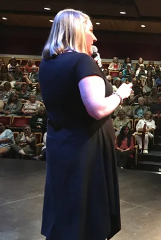 Nicole Powell with blonde hair speaking into a microphone on stage in front of an audience seated in an auditorium.