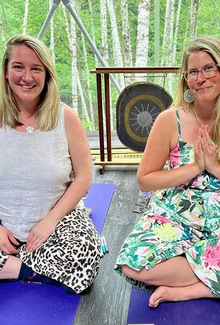 Two smiling women sitting cross-legged on yoga mats with a gong and birch trees in the background.
