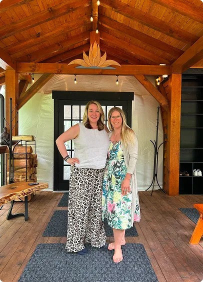 Two women smiling and standing arm-in-arm on a wooden porch with a high ceiling and decorative woodwork, one barefoot and wearing a floral dress, the other in leopard print pants and a white sleeveless top.
