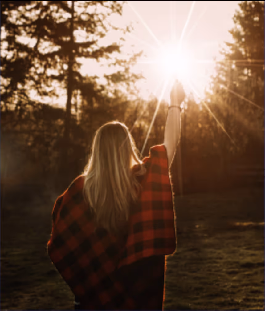 Person with long hair wearing a red and black plaid shirt reaching up towards the bright sun with trees in the background.