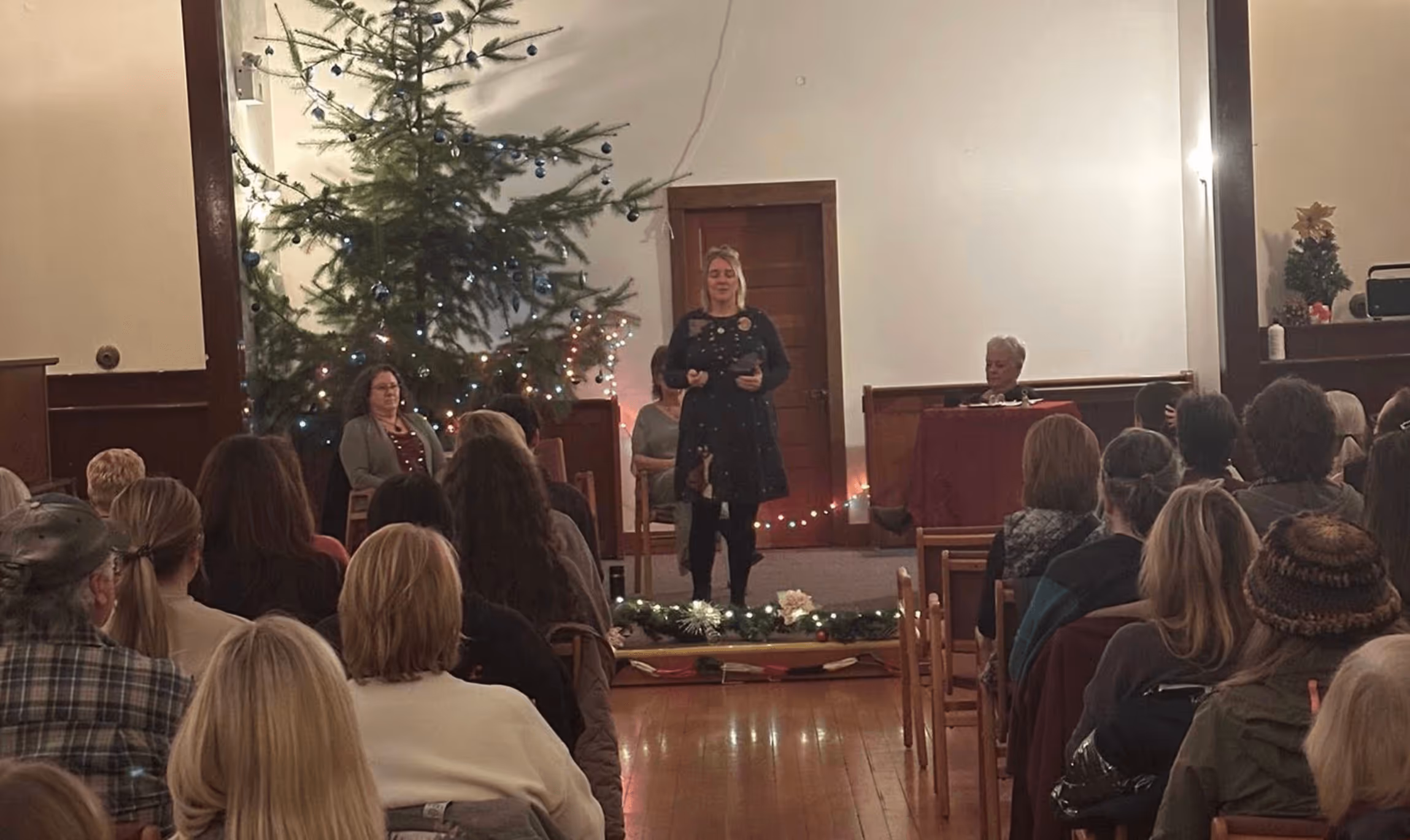 Woman in a black dress speaking to an audience in a room decorated with a Christmas tree and holiday lights.