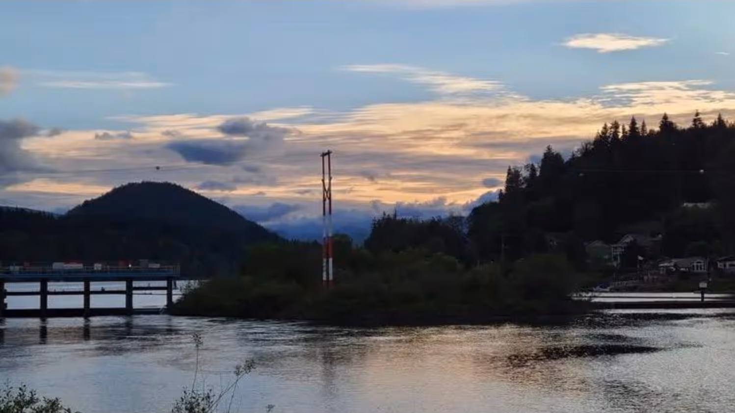Calm river at sunset with hills, trees, and a bridge in the background.
