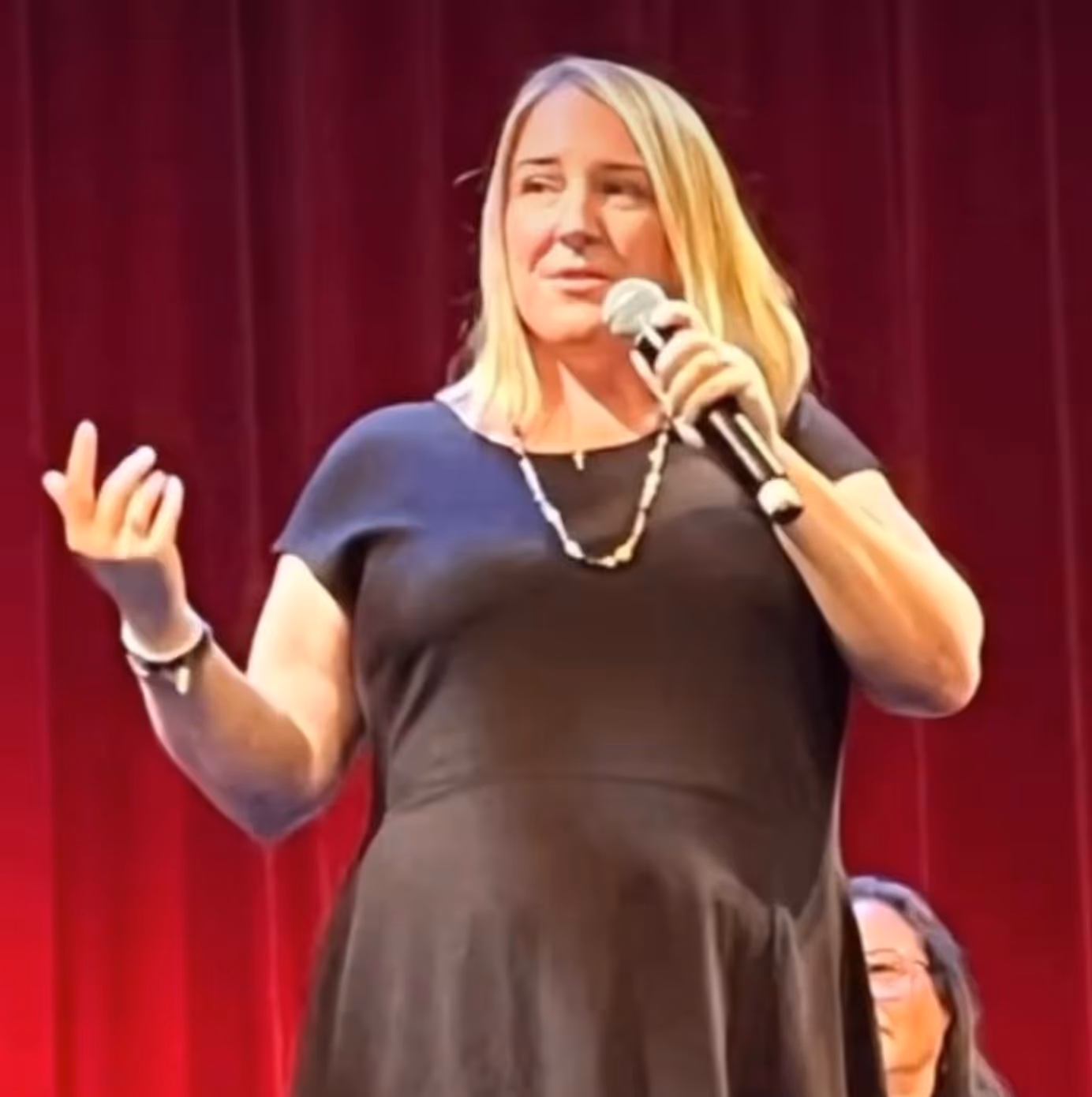 Nicole Powell speaking into a microphone on stage with a red curtain background.