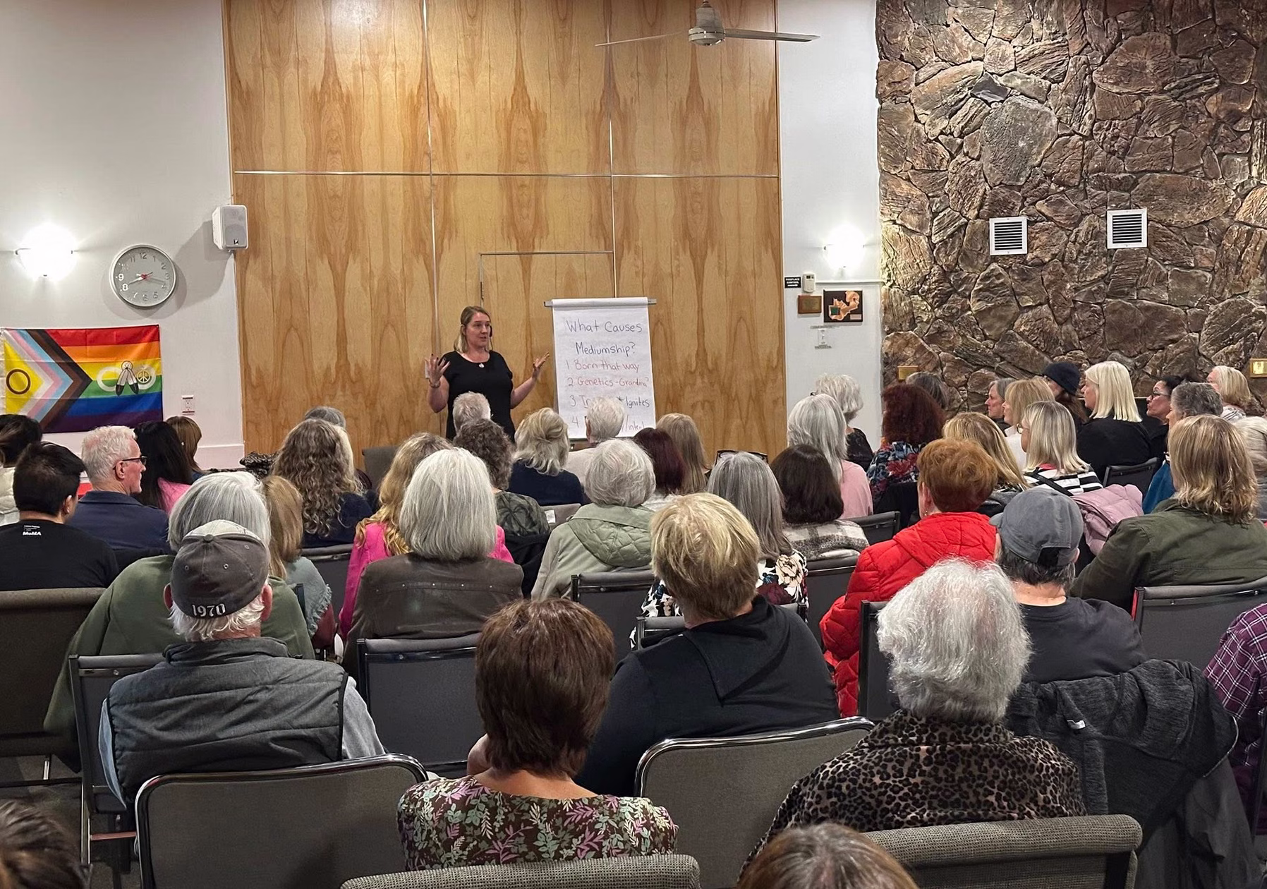 Nicole Powell Spiritual Medium speaking to an audience seated in rows inside a room with wooden paneling and a stone wall, next to a flip chart listing causes of mediumship.