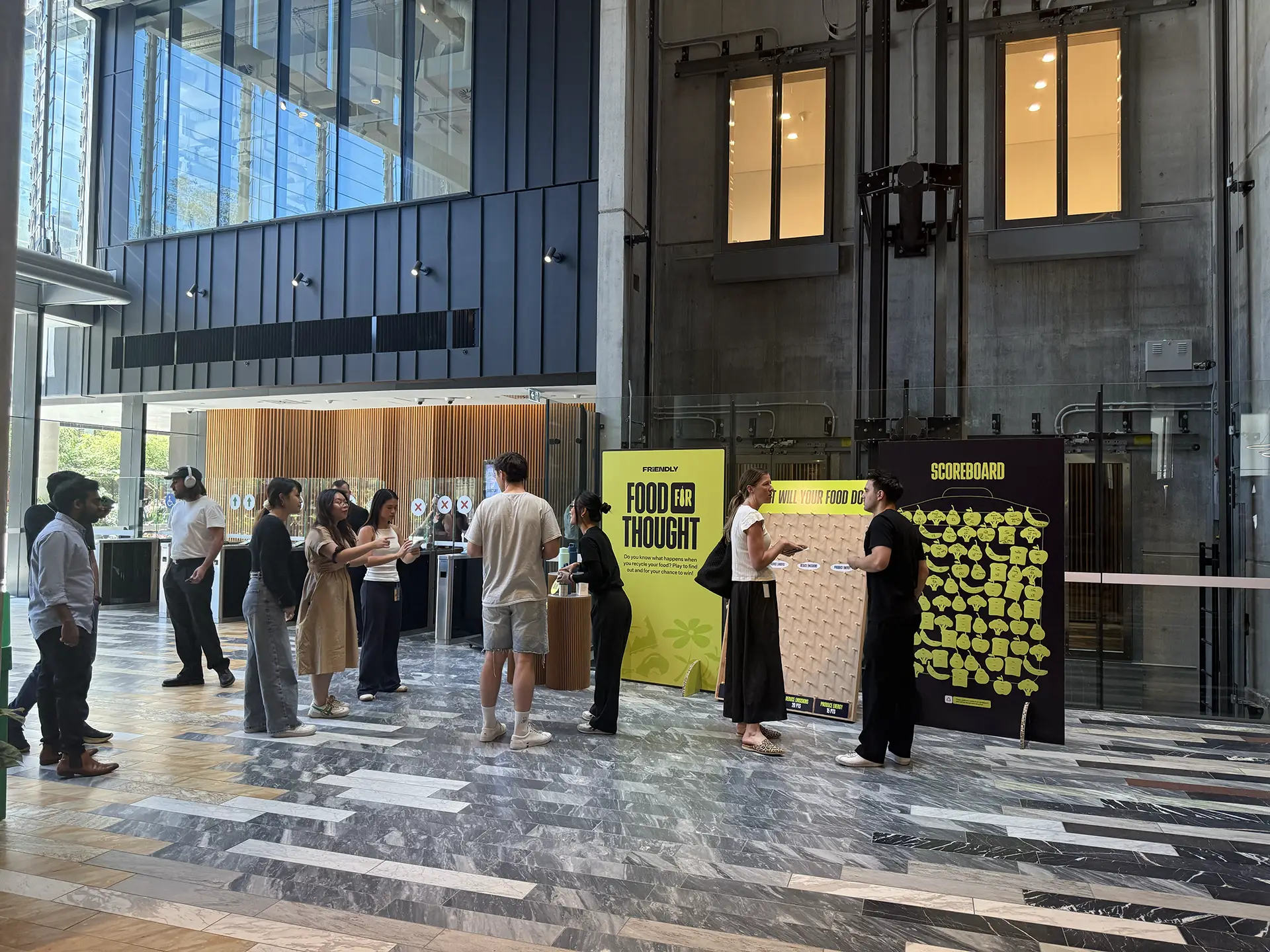 Office workers participating in a Friendly “Food for Thought” waste-reduction activation, interacting with large engagement boards in a building lobby. 