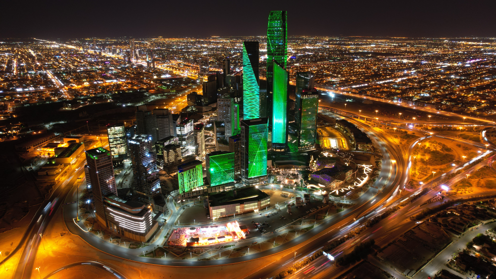 Night aerial view of Riyadh with illuminated skyscrapers lit in green and surrounding highways bustling with traffic.