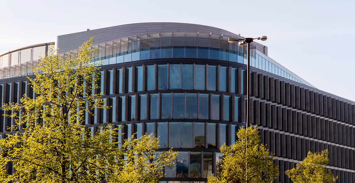 Modern office building with curved glass facade partially obscured by green trees and a streetlight in front.