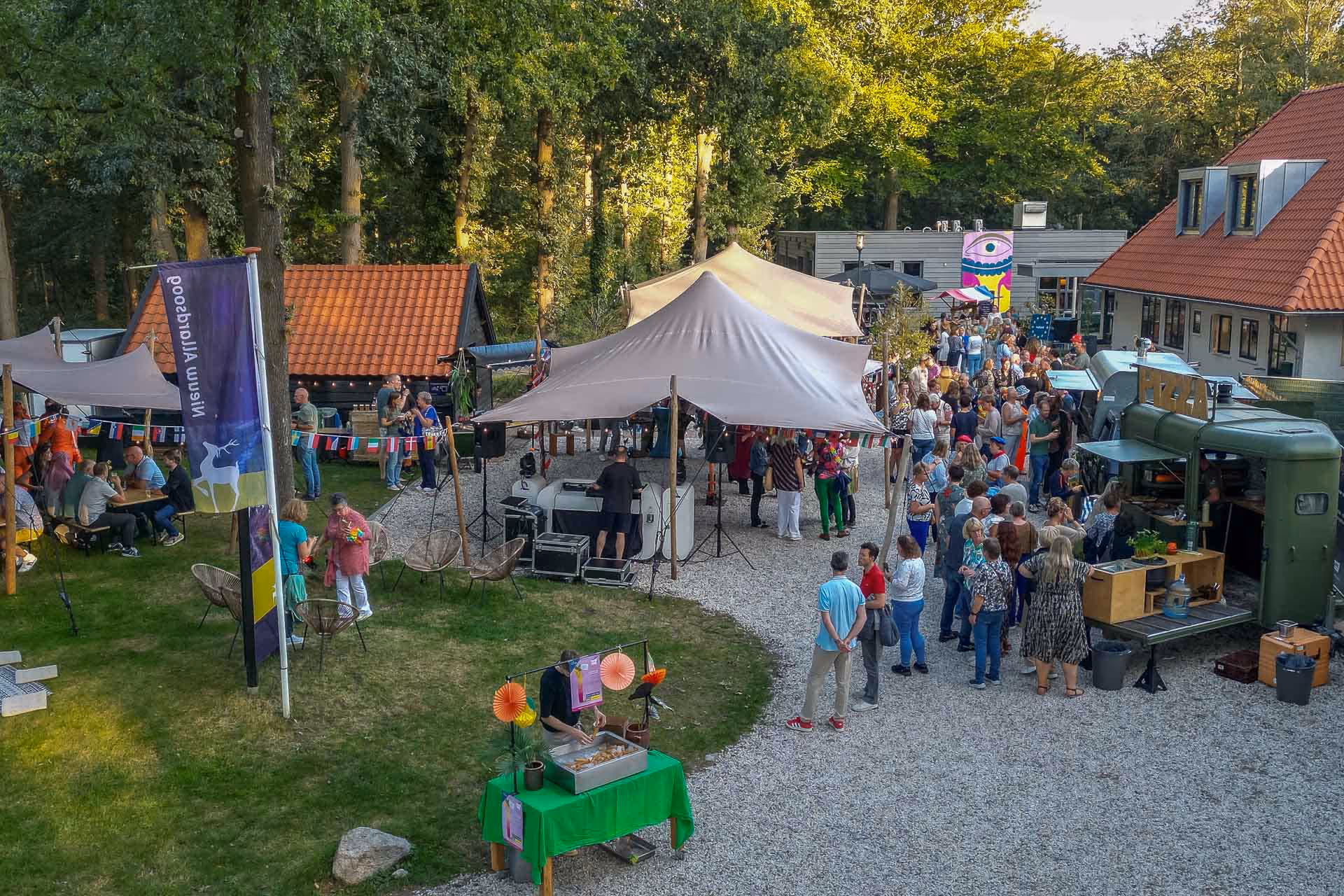 Een drukbezocht buitenfeest in een park met een tent, een pizzakraam, en mensen die in de rij staan en sociale gesprekken voeren.