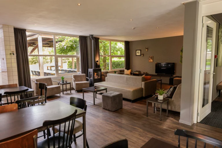 Gezellige woonkamer met grote ramen, banken, fauteuils, een houtkachel en een tv aan de muur.