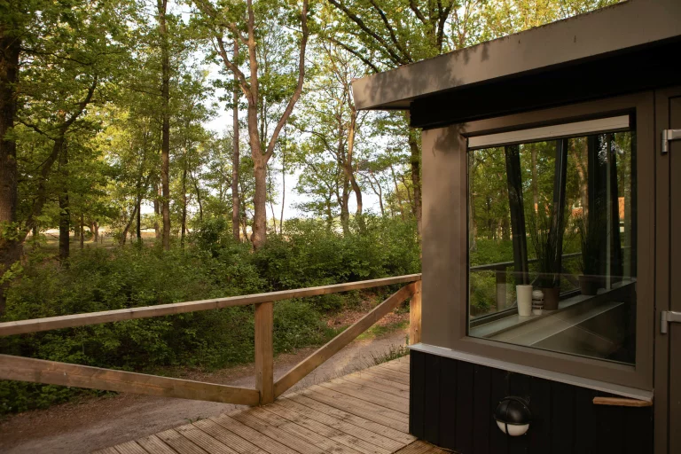 Houten terras met houten hekwerk naast een gebouw met een grote raam in een bosrijke omgeving.
