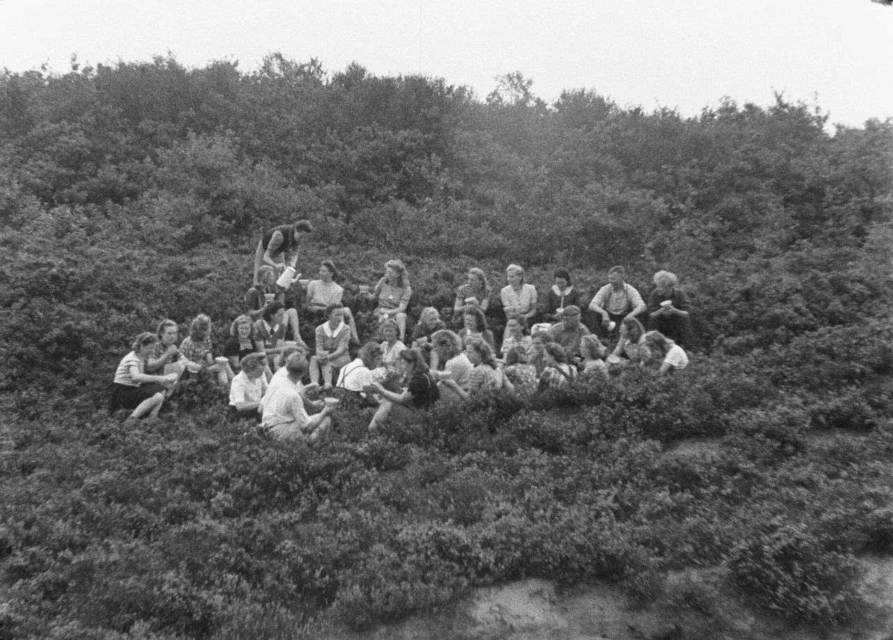Groep jongeren zittend op grasheuvel in buitengebied, sommigen praten en lachen, zwart-wit foto.