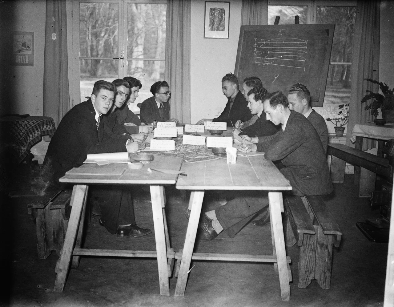 Tien mannen zitten aan een tafel met papieren en naamkaartjes, mogelijk tijdens een vergadering of klas, naast een schoolbord met grafieken.