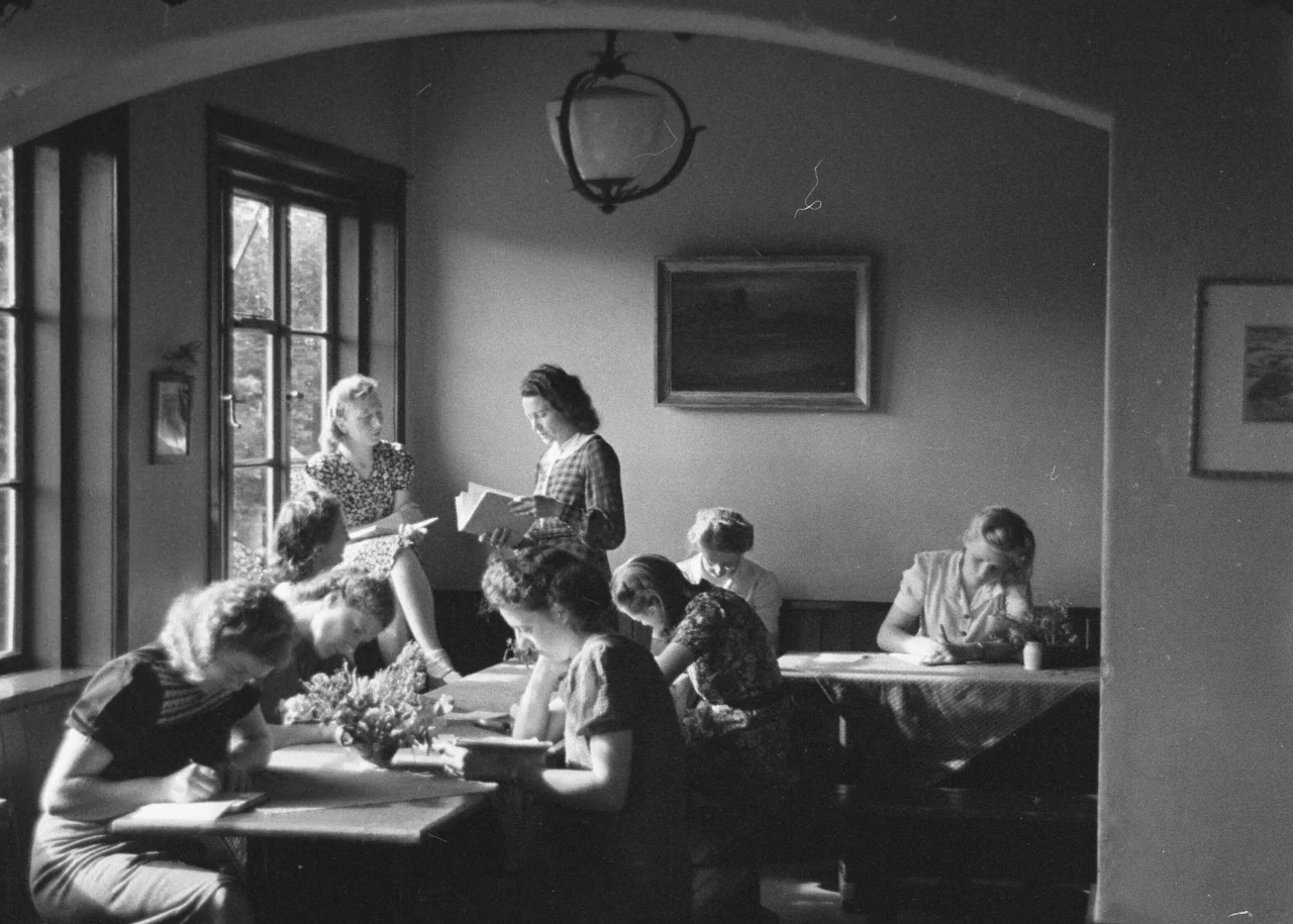 Een zwart-witfoto van een groep vrouwen die aan tafels zitten en lezen of schrijven in een kamer met grote ramen en eenvoudige inrichting.