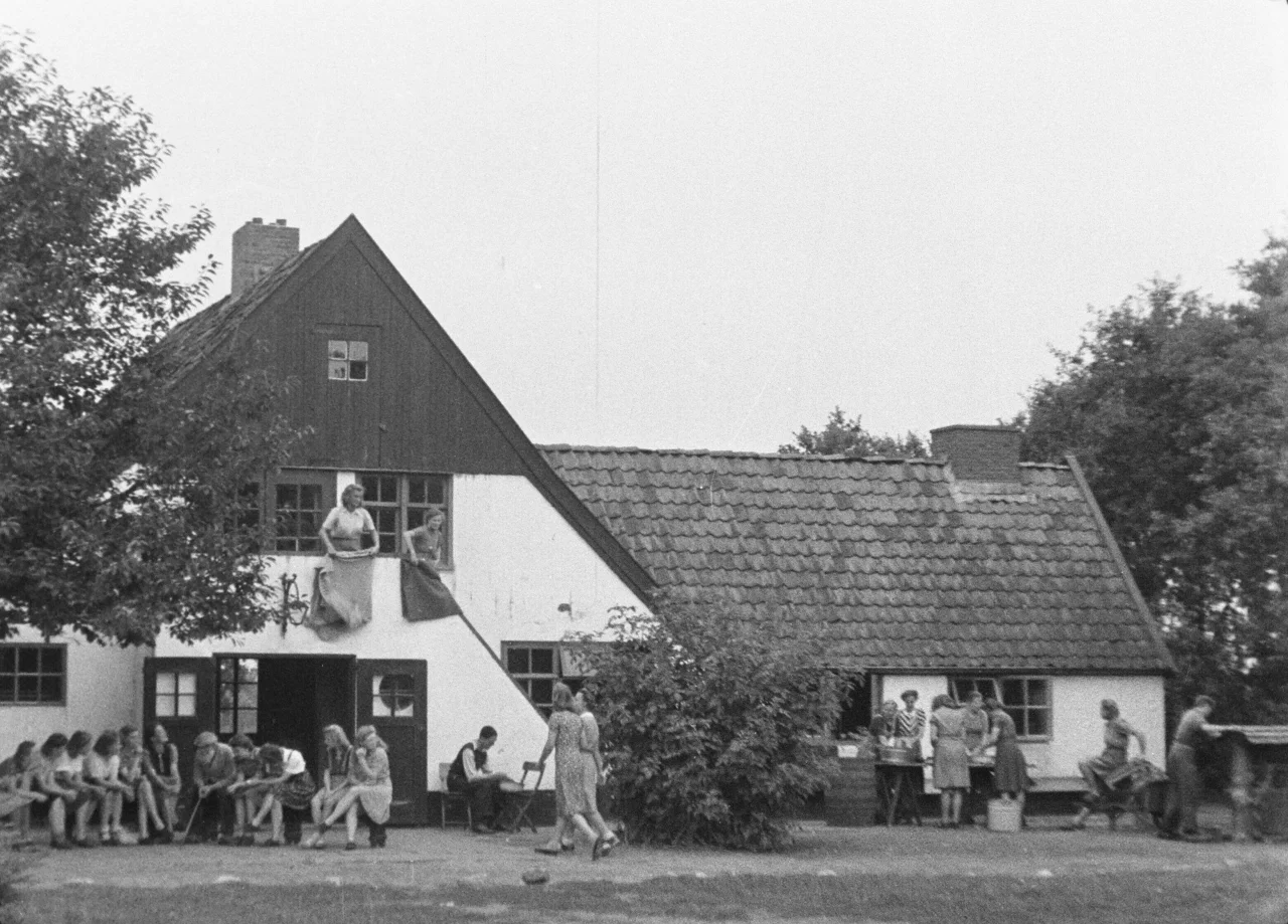 Zwart-wit foto van een groep mensen bij een traditioneel huis met schilddak en houten kozijnen, sommigen zitten op een bank, anderen staan of hangen uit het raam.