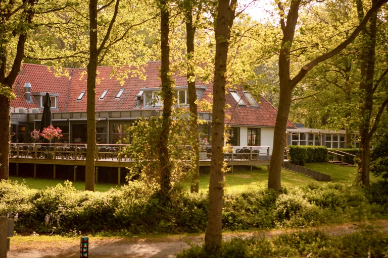 Gezellig terras van een restaurant met rode dakpannen, omgeven door groene bomen en zonlicht.