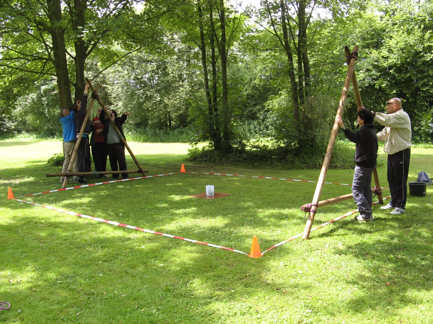Groep mensen werkt samen met houten stokken binnen een omheind vierkant grasveld in een groene parkachtige omgeving.