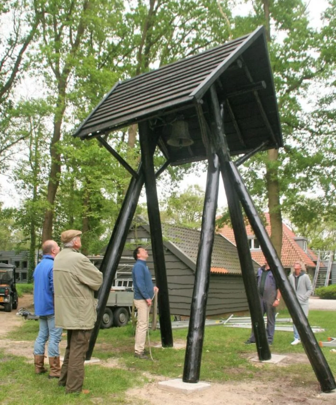 Vijf mannen kijken naar een grote klok die aan een houten constructie met een zwart dak hangt in een groene omgeving met bomen en gebouwen.