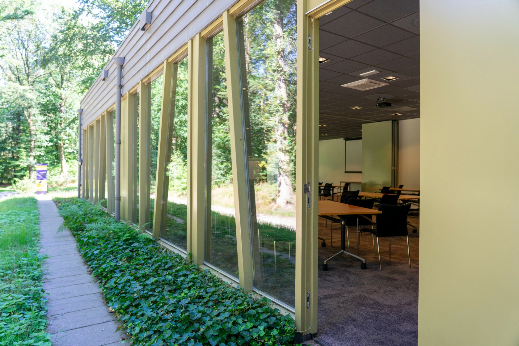 Gebouw met grote ramen naast een groen pad en gedeeltelijk zicht op een klaslokaal met tafels en stoelen.