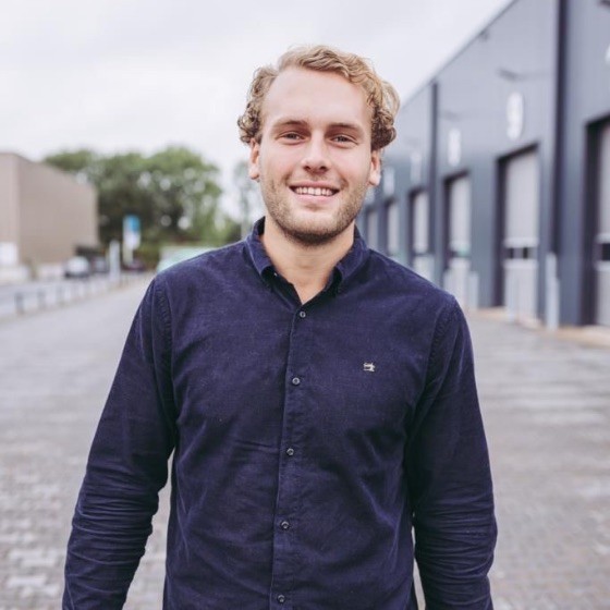 Jonge man met blonde krullen en een donkerblauw overhemd staat buiten op een geplaveide straat met moderne industriële gebouwen op de achtergrond.