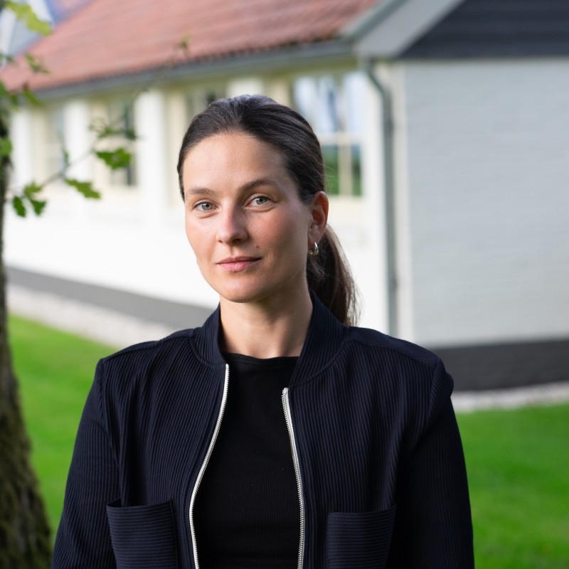 Vrouw met zwarte jas en donkere haren in een paardenstaart, staand in de tuin voor een wit huis met rood dak.