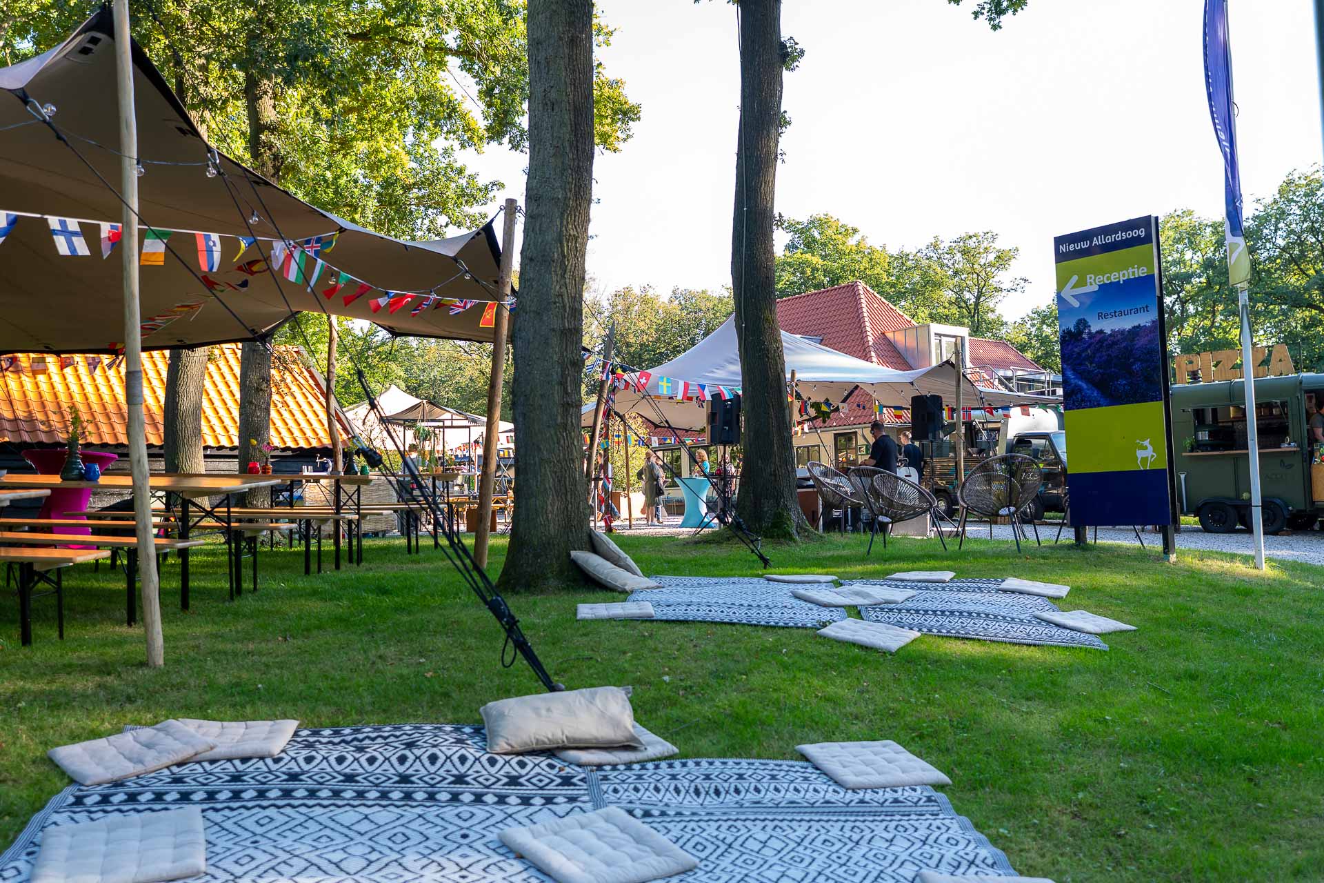 Outdoor event area with picnic tables, patterned rugs with cushions on grass, tents with international flags, and a sign pointing to reception and restaurant.