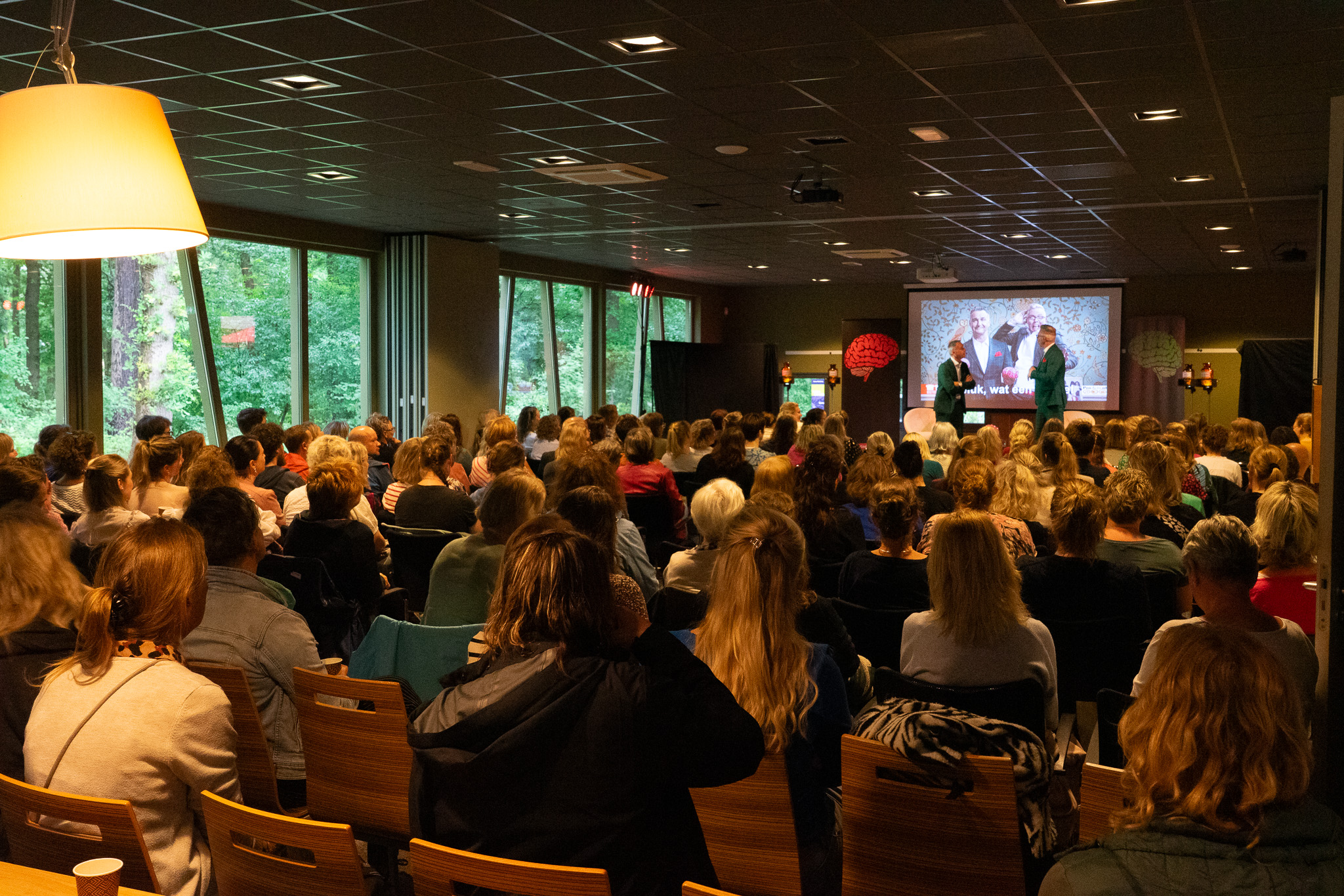 Grote groep mensen zit in een verduisterde zaal en kijkt naar een presentatie met twee sprekers voor een scherm.