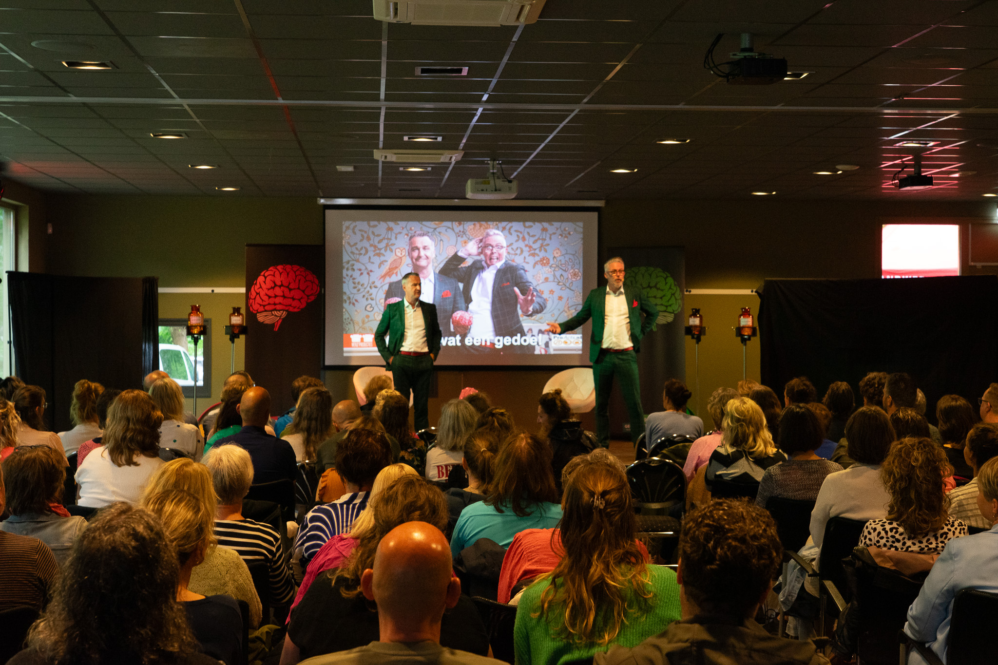 Twee mannen in groene pakken presenteren voor een zaal vol mensen met een projectie van twee mannen en hersenen op het scherm achter hen.