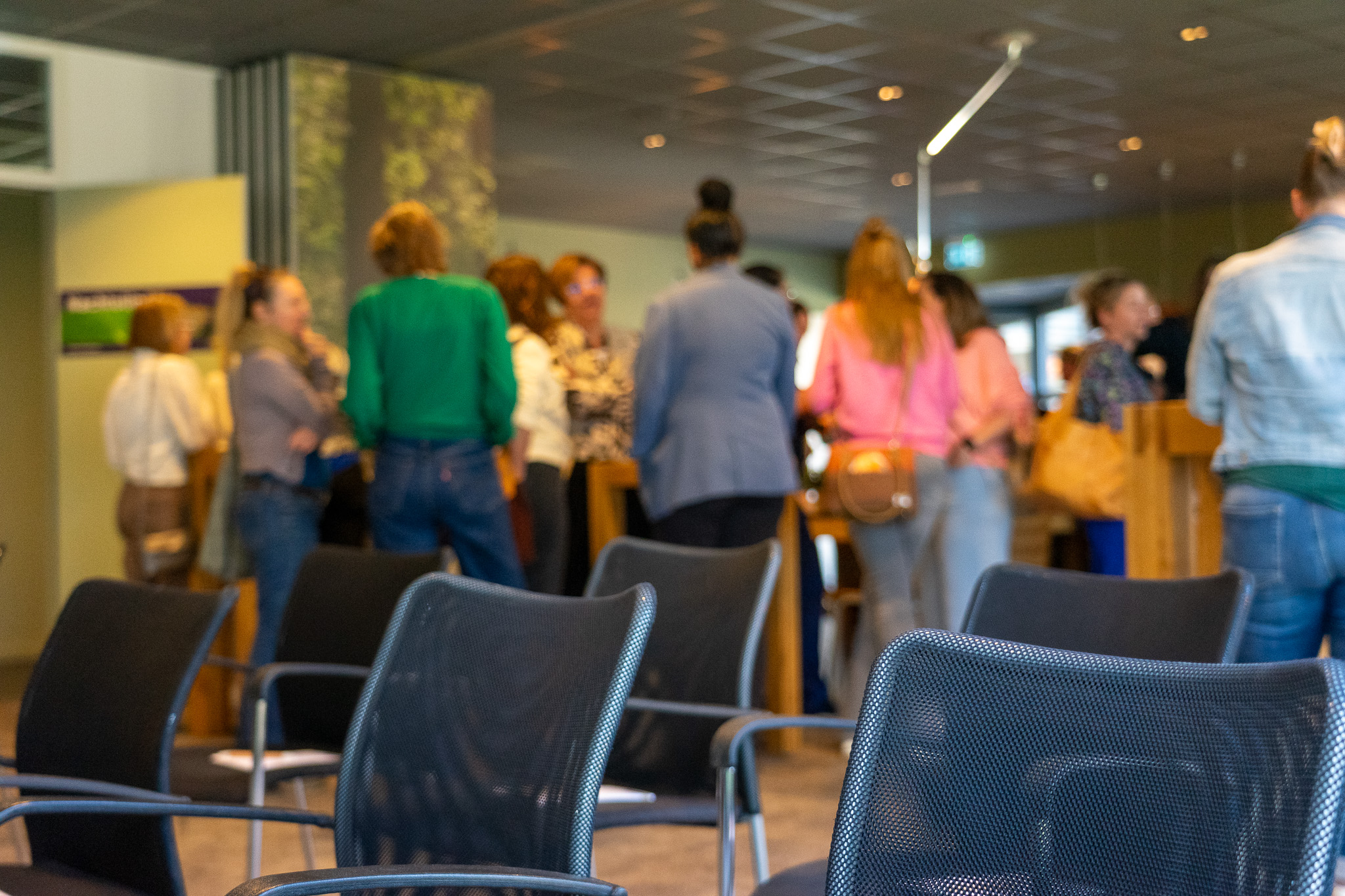 Groep mensen die in een binnenruimte staan en gesprekken voeren, met lege stoelen op de voorgrond.