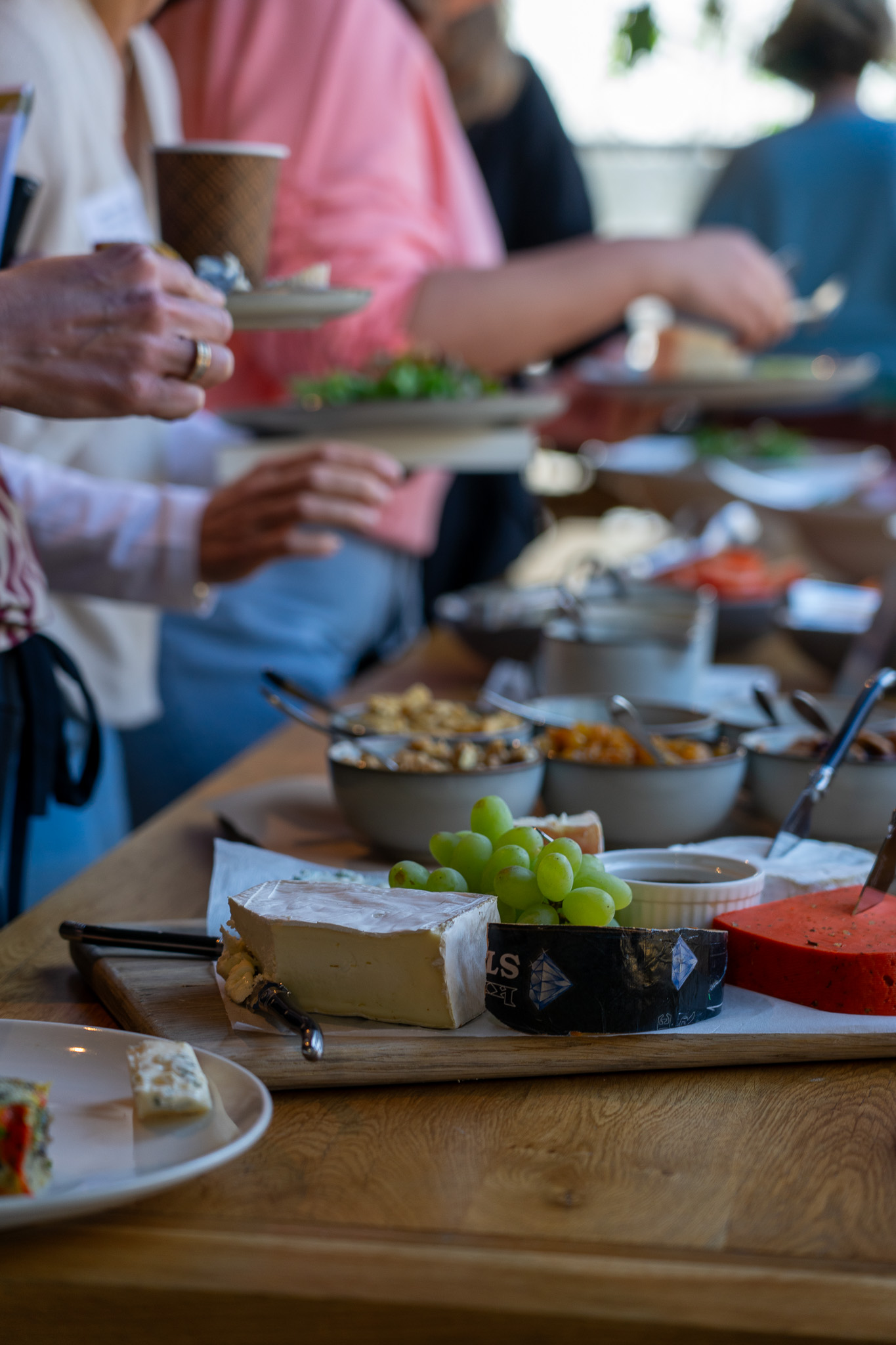 Plank met verschillende kazen, druiven en eetlepels met spreads op een houten tafel bij een buffet.