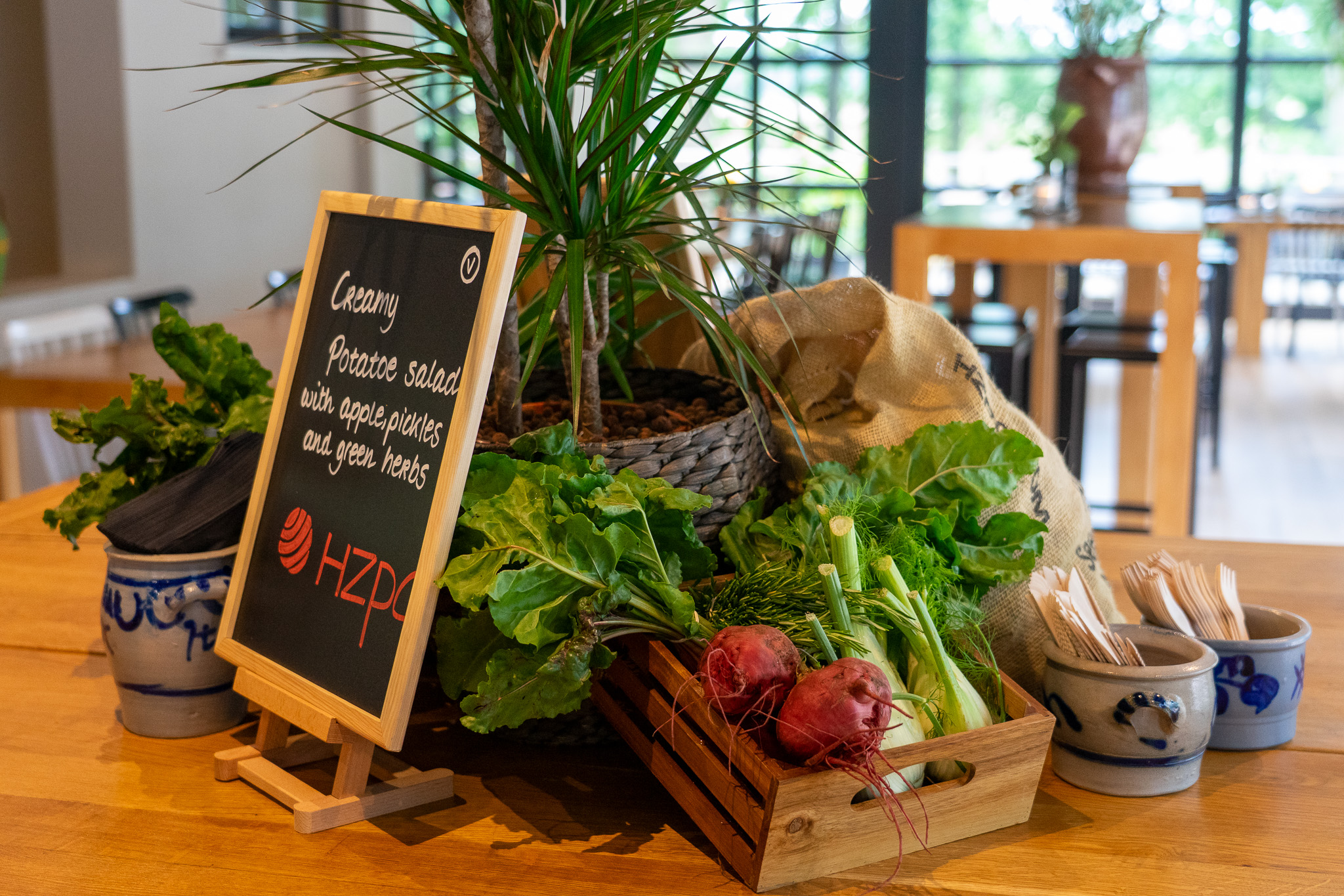 Een houten tafel met een houten krat met verse groenten zoals bieten en kruiden, een bordje met het menu voor romige aardappelsalade en bestekhouders.