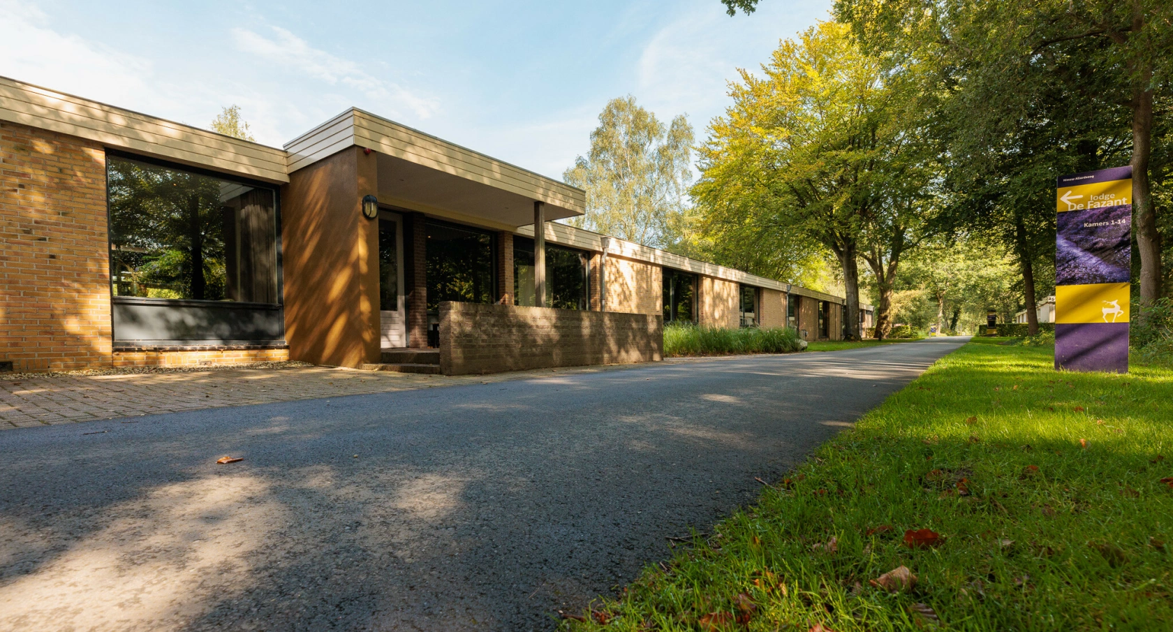 Lang gebouw van rode baksteen met grote ramen langs een met asfalt bestraatte weg, omringd door groen en bomen in zonlicht.