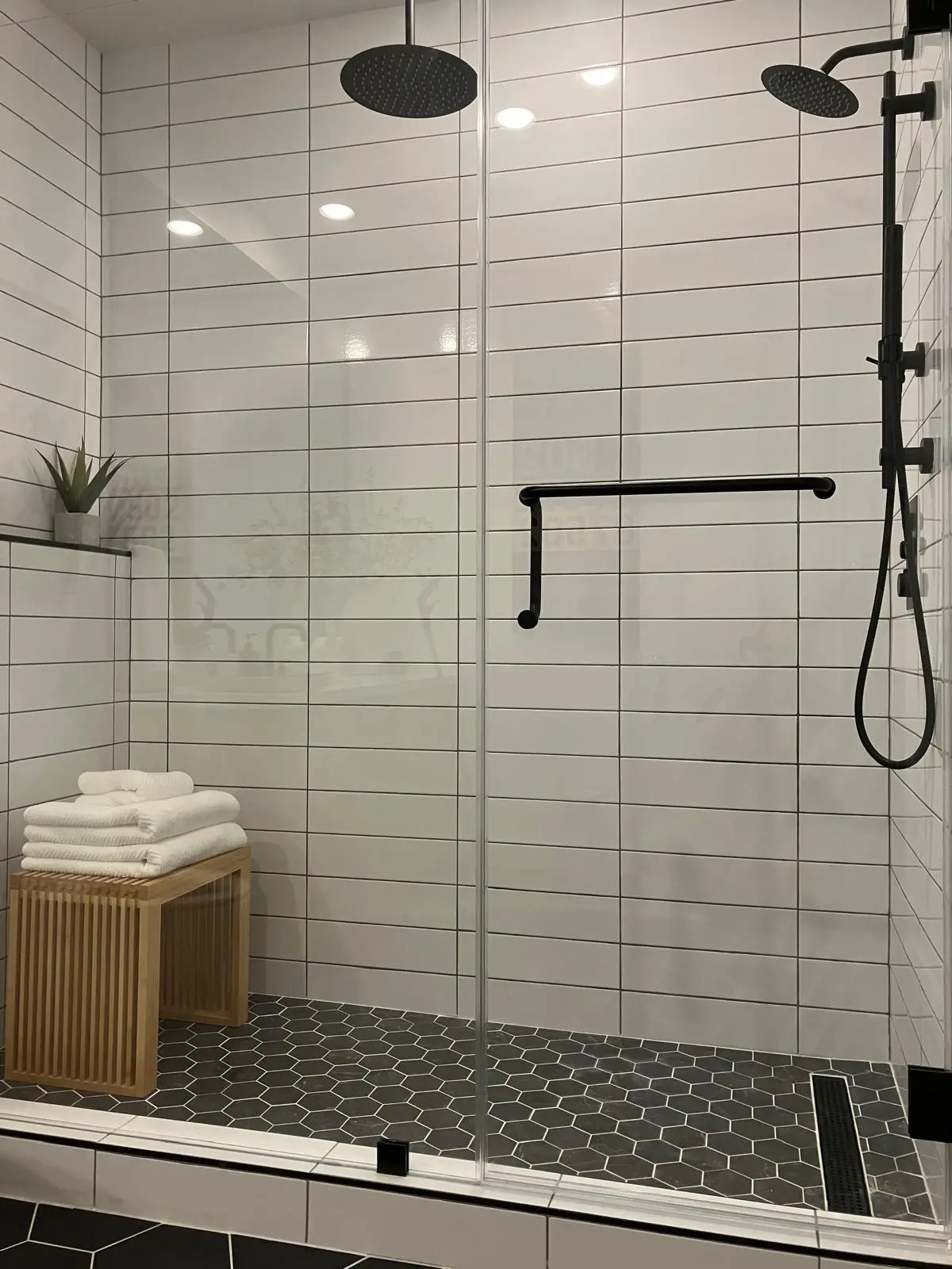 Modern bathroom shower with white subway tile walls, black hexagonal floor tiles, glass door, and wooden bench with towels