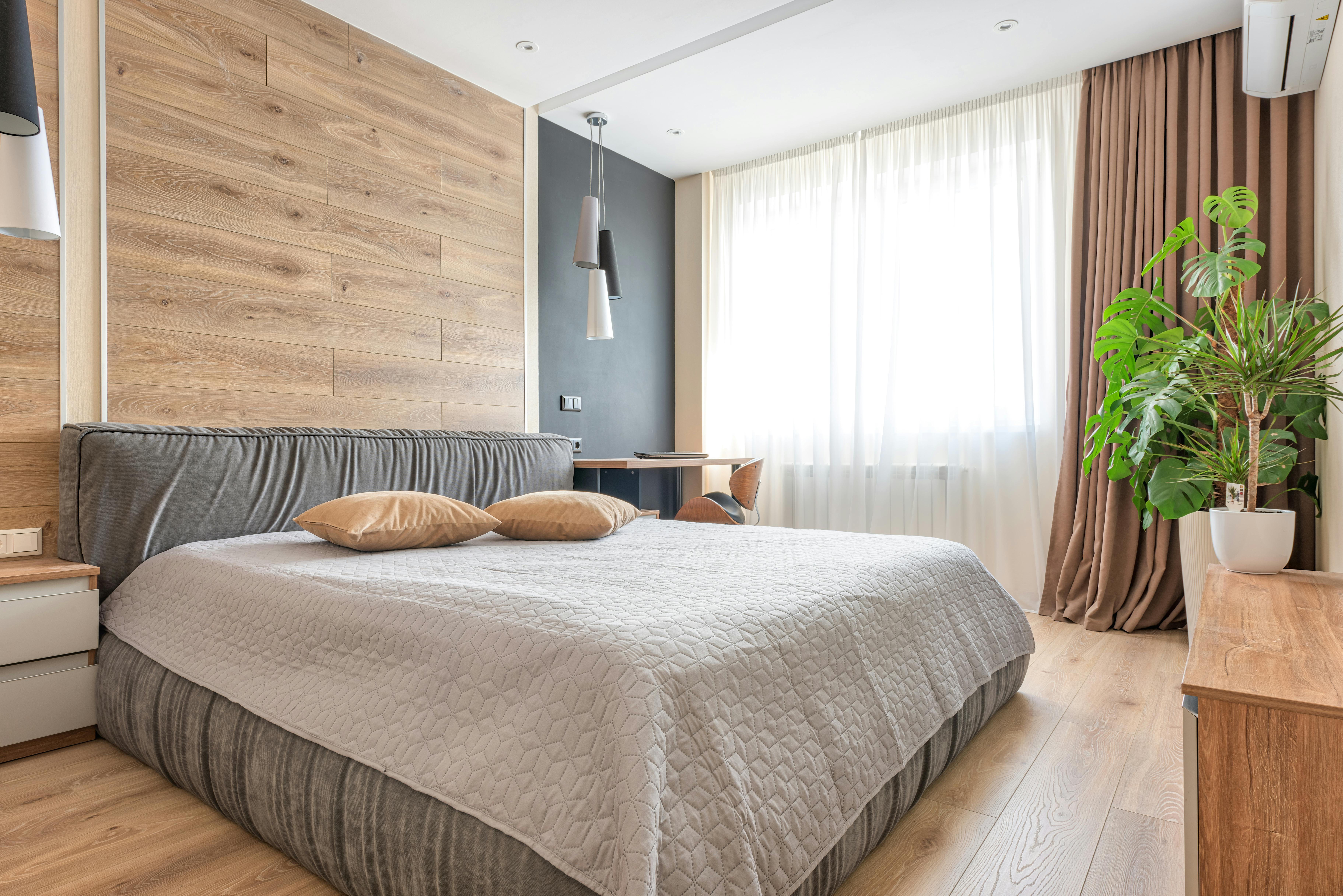 Modern bedroom with wood accent wall, gray upholstered bed, white quilted bedding, pendant lights, and large window with natural light