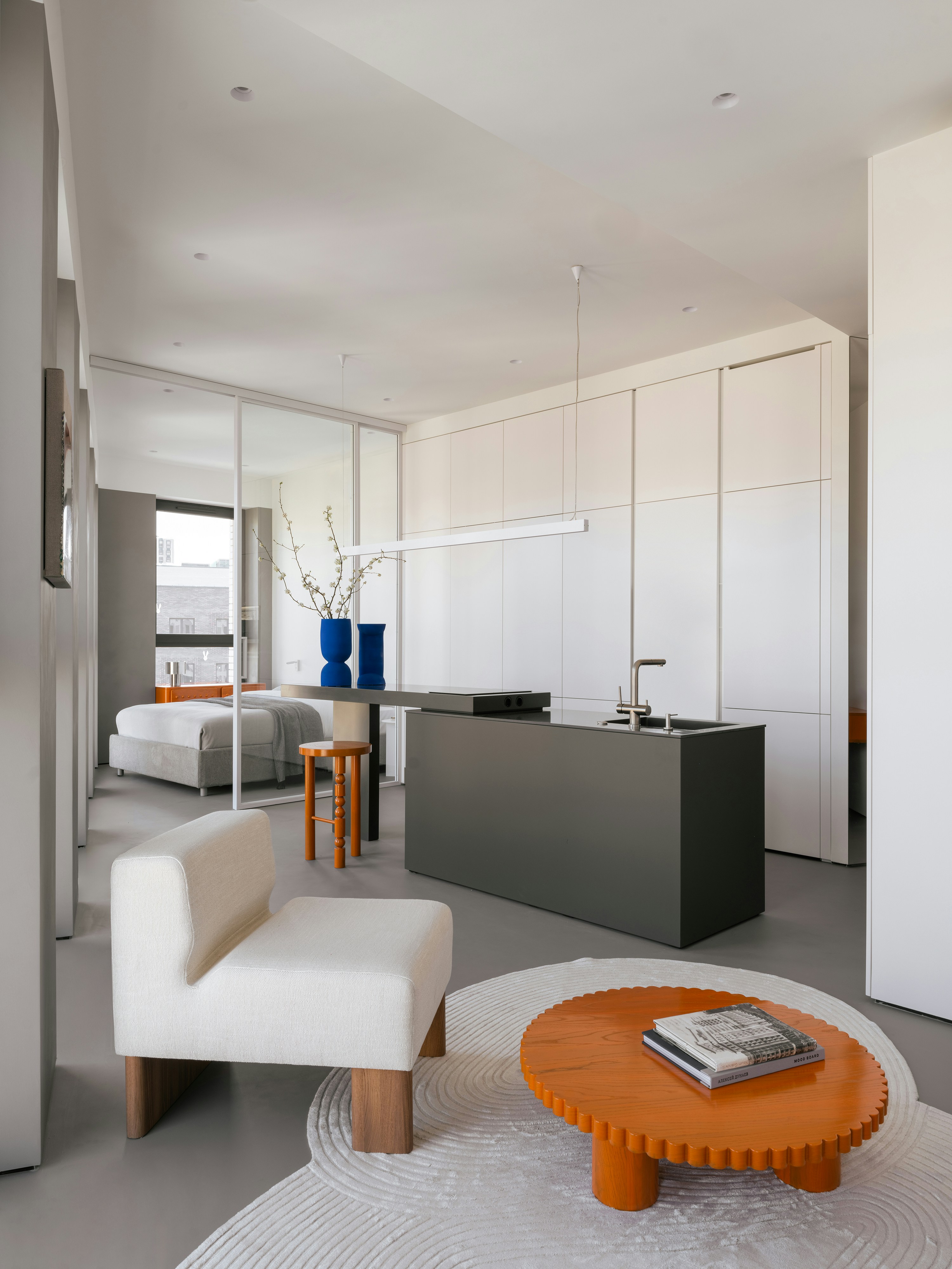 Modern studio apartment with white kitchen cabinets, dark green island, cream upholstered chair, and orange round coffee table