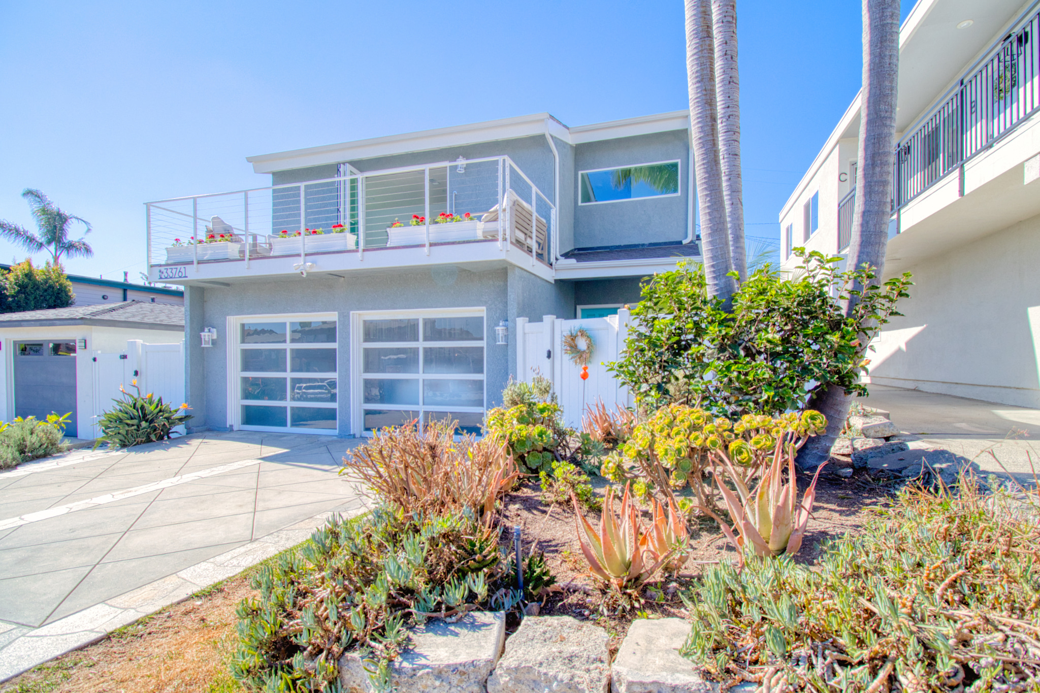 Modern two-story coastal home with gray siding, glass garage doors, white railings, and drought-resistant landscaping