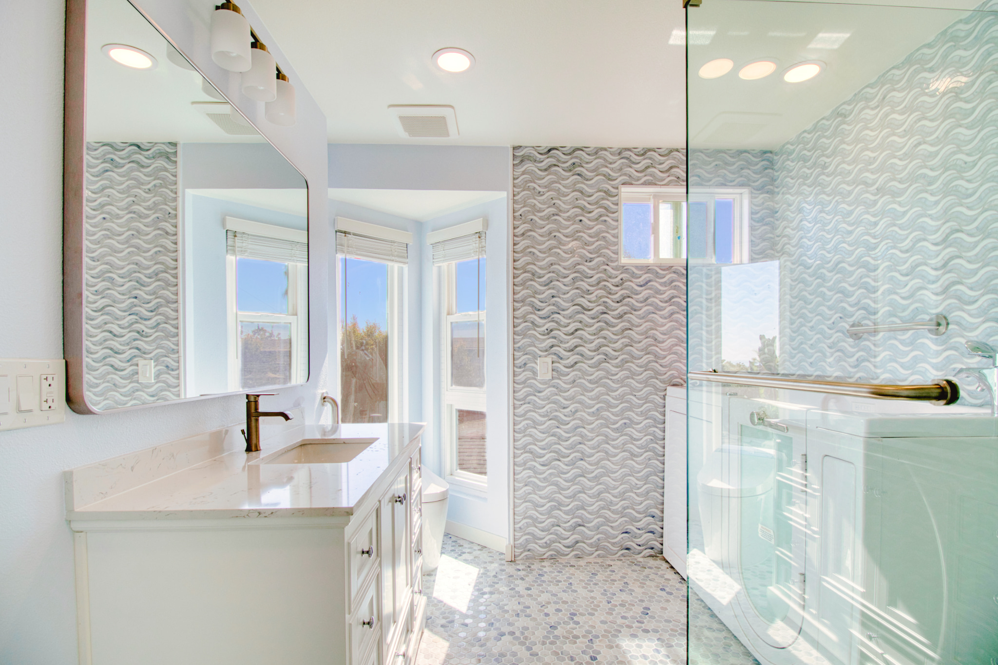 Modern bathroom with white vanity, brass fixtures, blue-gray wave tile walls, glass shower, and natural light from windows