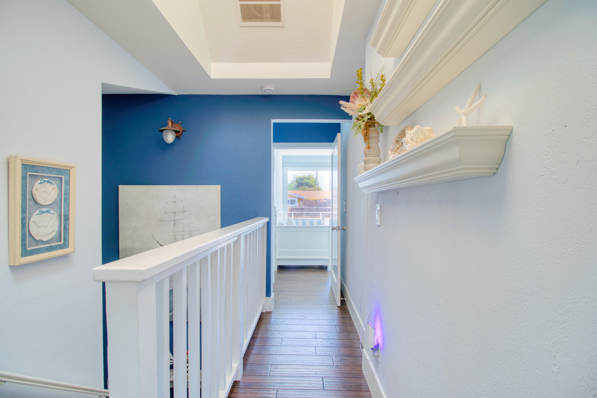 Upstairs hallway with white railings, blue accent wall, nautical artwork, crown molding, and dark hardwood floors