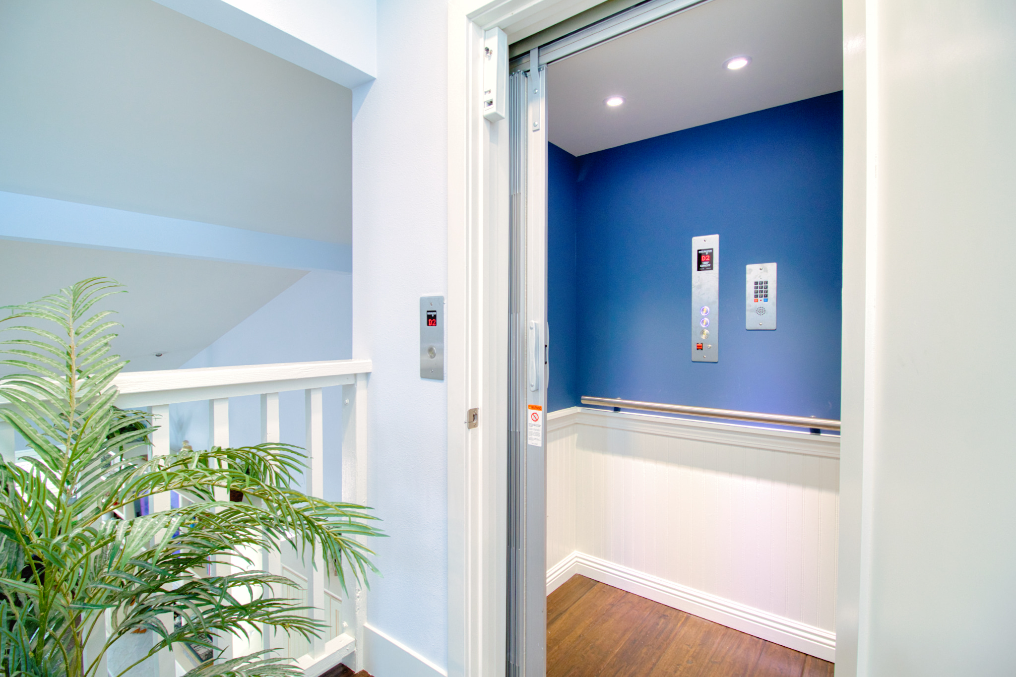 Modern elevator with blue accent wall and control panels, adjacent to white staircase railing with decorative palm plant