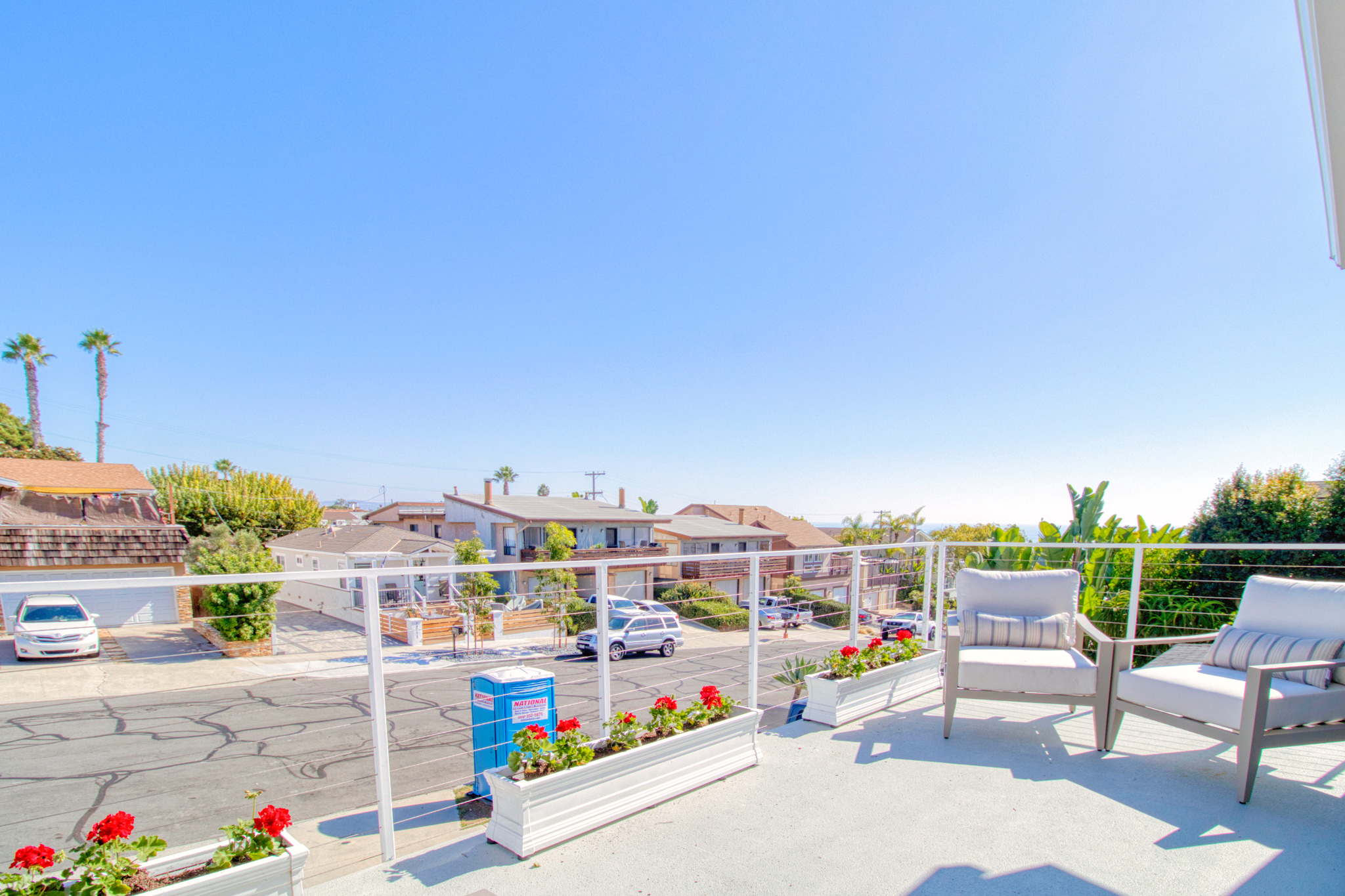 Modern rooftop deck with white cable railing, gray outdoor chairs, red geraniums in planters, overlooking residential neighborhood