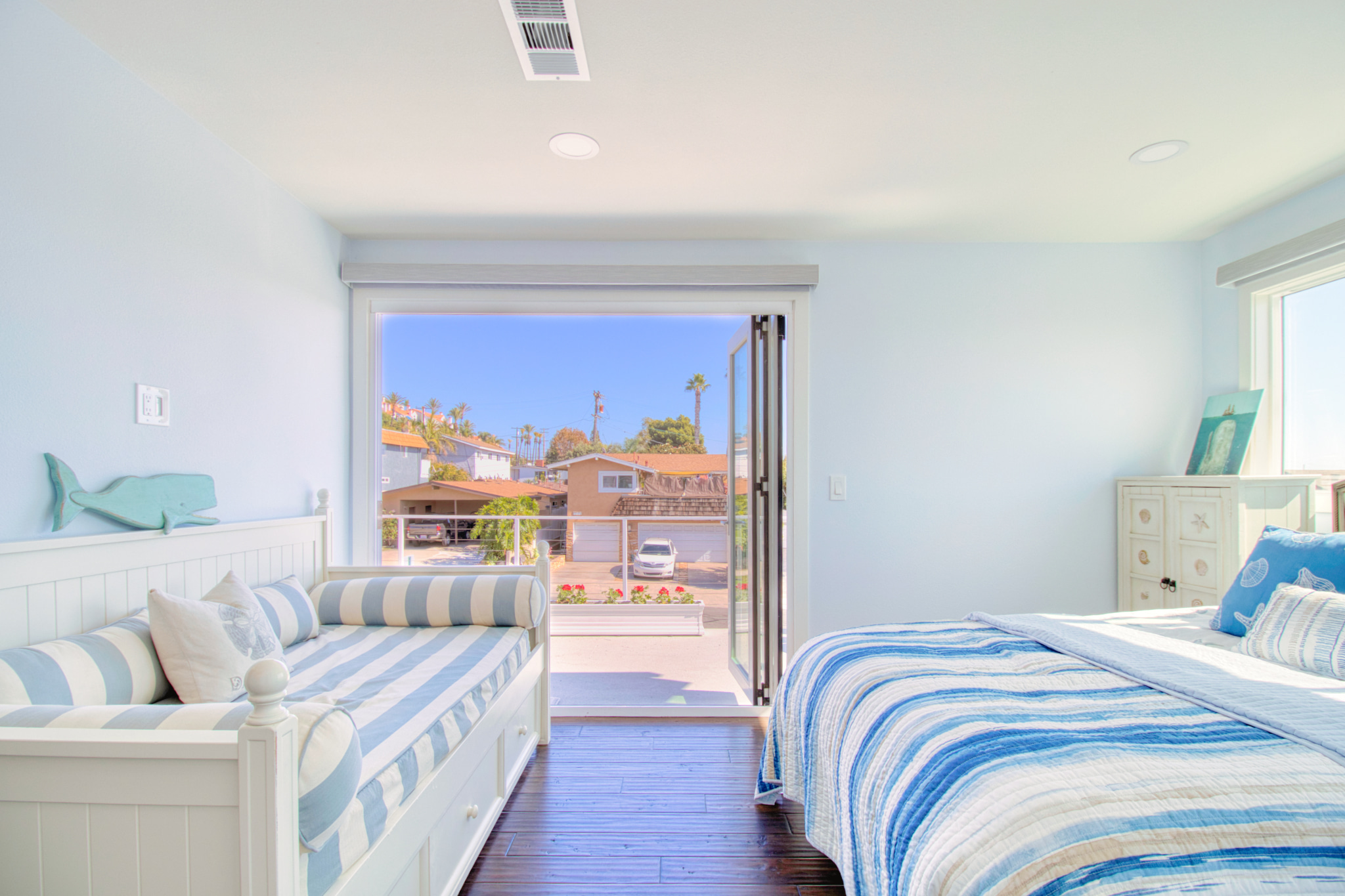 Coastal-style bedroom with blue and white striped bedding, white furniture, and sliding door opening to sunny California neighborhood