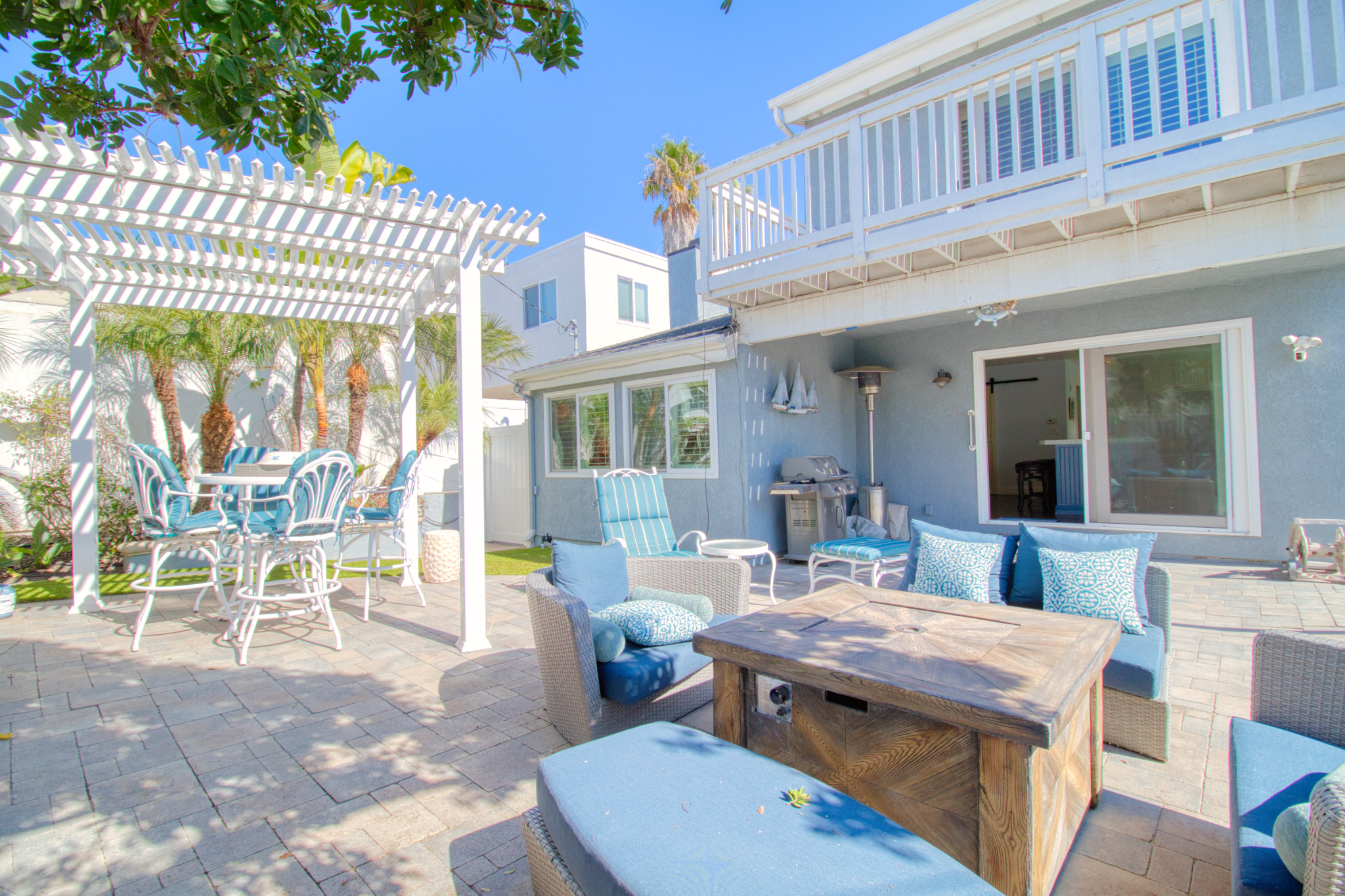 Coastal outdoor patio with blue cushioned wicker furniture, rustic wood coffee table, white pergola, and stone pavers