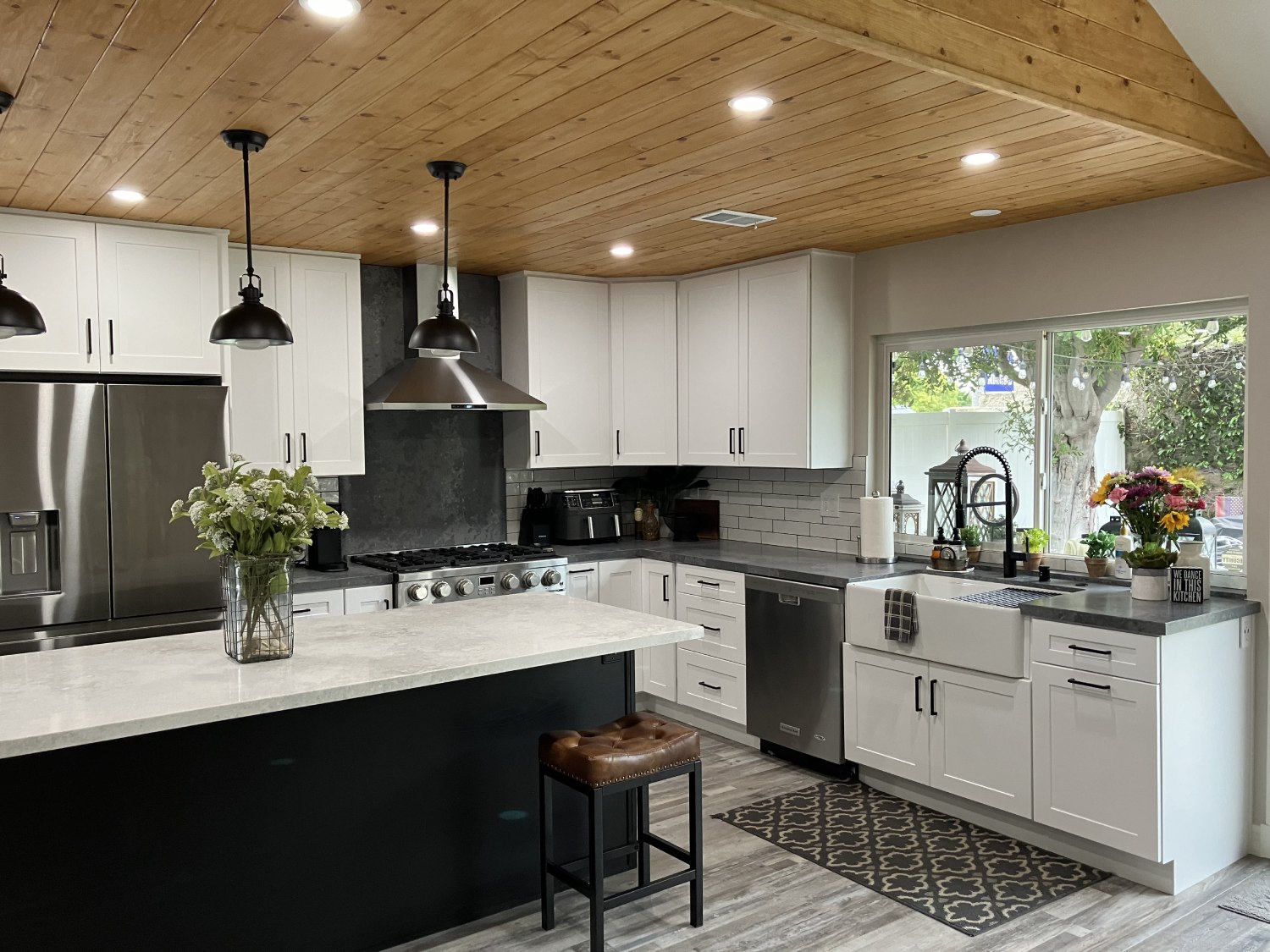 Modern farmhouse kitchen with white shaker cabinets, dark countertops, wood plank ceiling, subway tile backsplash, and black pendant lights
