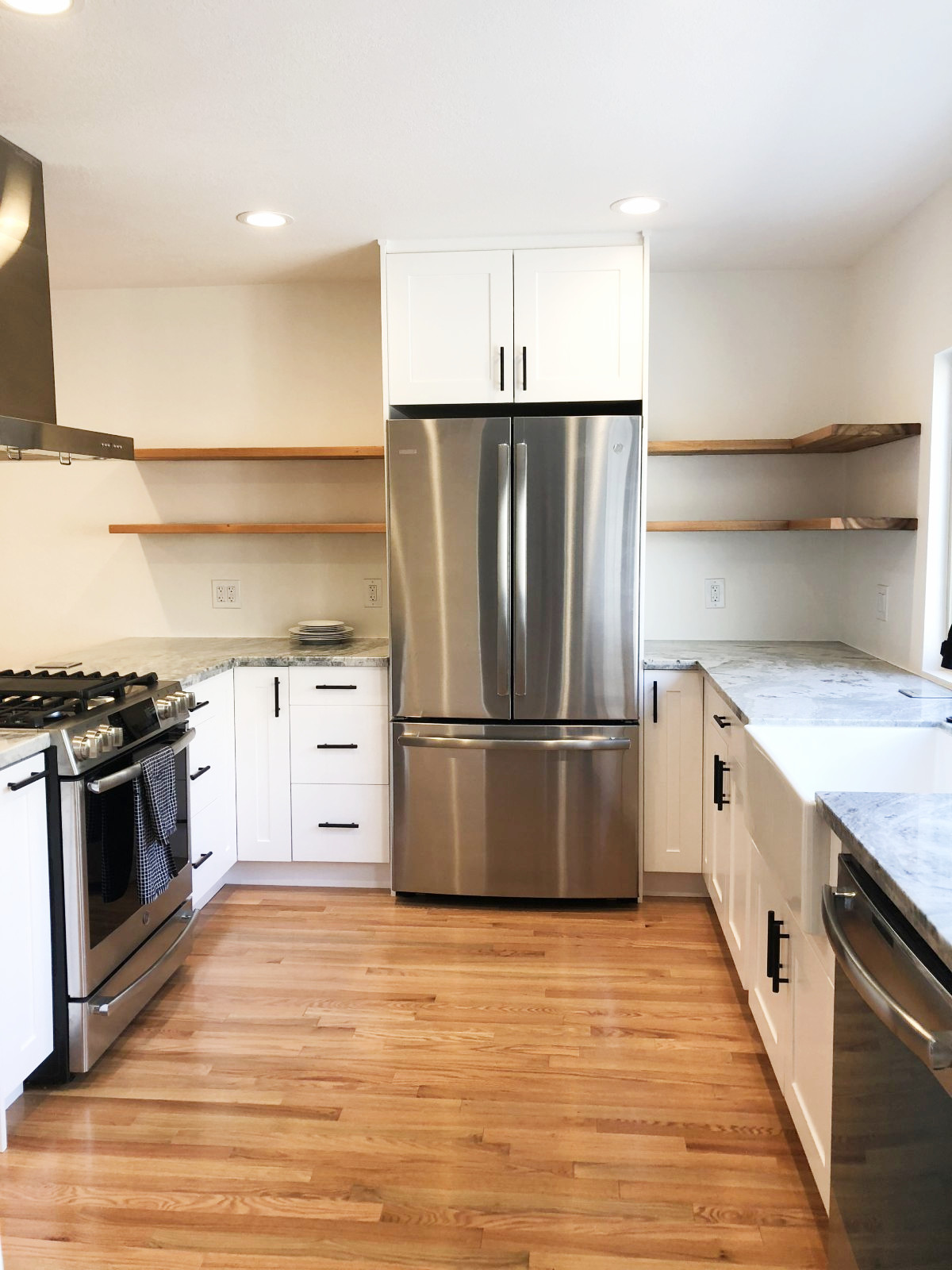 Modern white kitchen with stainless steel appliances, marble countertops, wood floating shelves, and hardwood floors