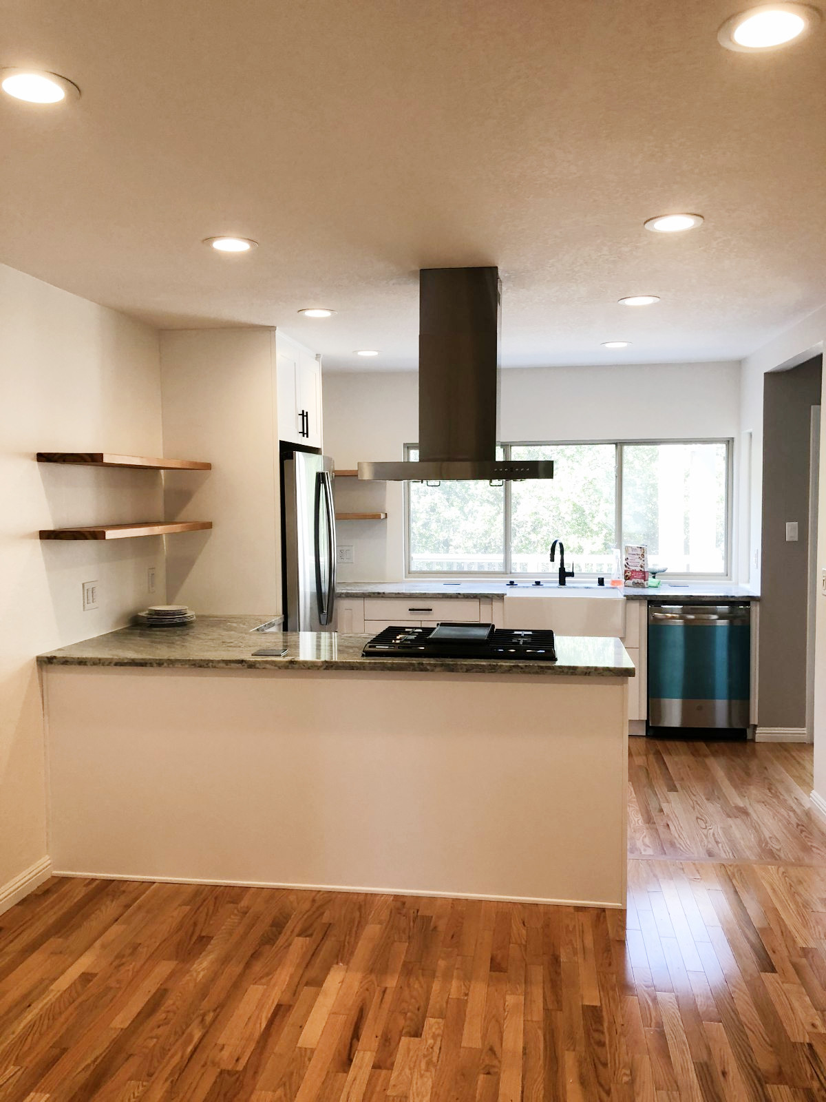 Modern white kitchen with granite island, black cooktop, stainless steel appliances, wood shelving, and hardwood floors