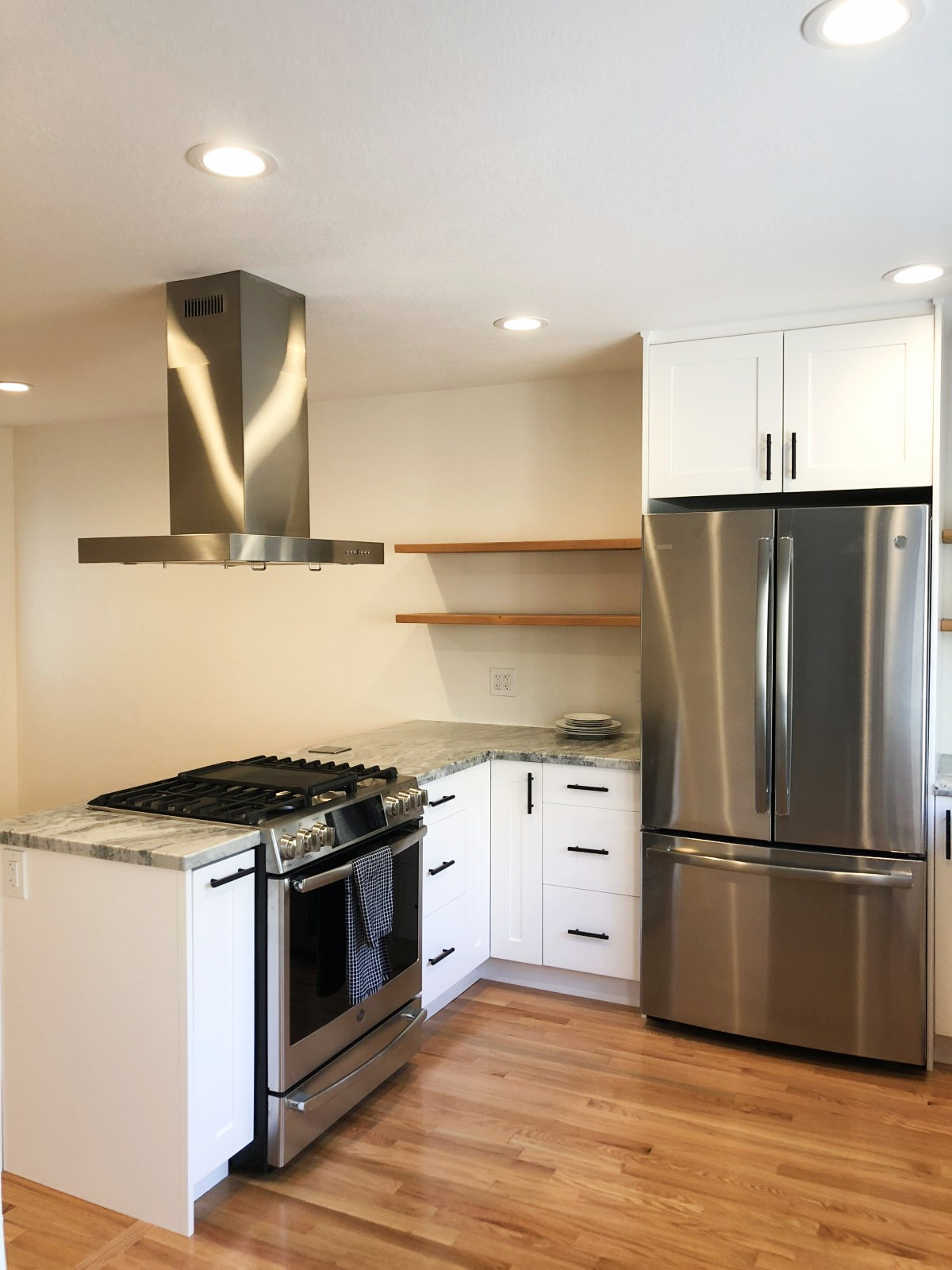Modern white kitchen with stainless steel appliances, granite countertops, floating wood shelves, and hardwood floors