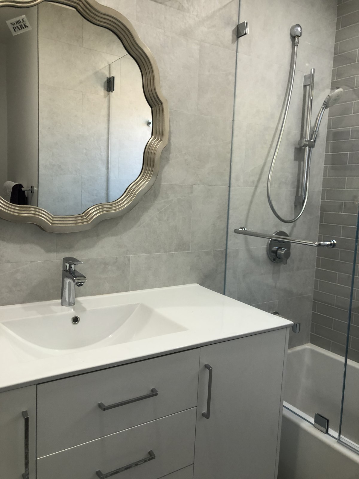 Modern bathroom with white vanity, decorative wavy mirror, glass shower door, and gray tile walls in neutral tones