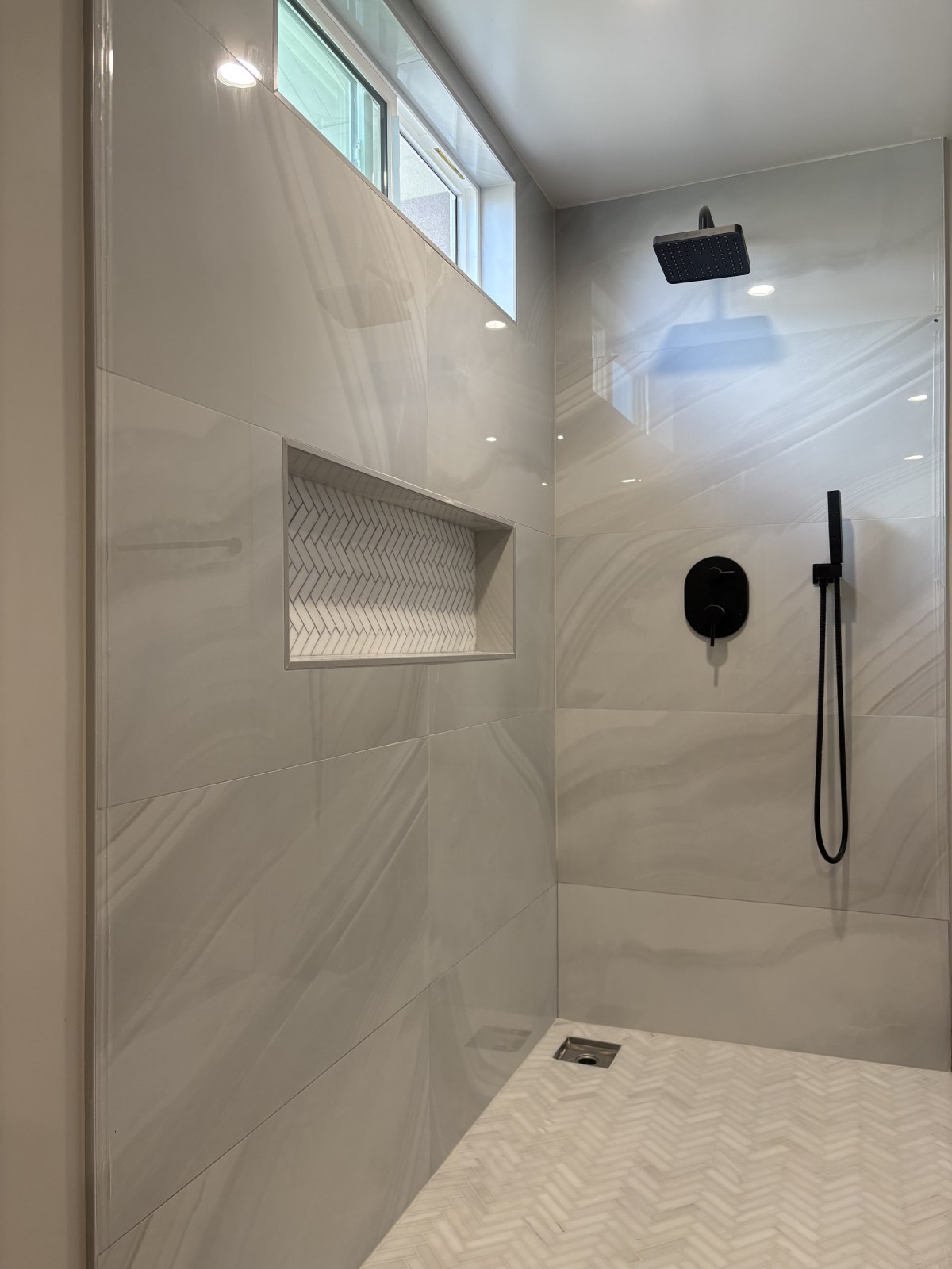 Modern walk-in shower with large white marble-look tiles, built-in niche with herringbone accent, black fixtures and rainfall showerhead